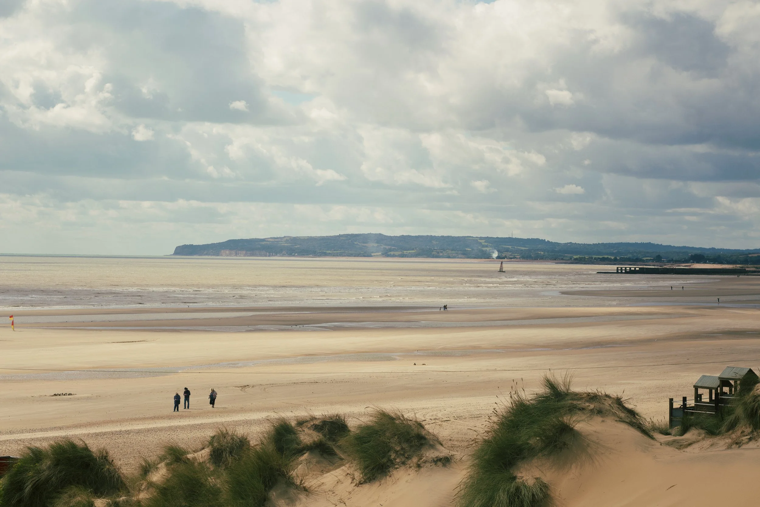 A beach with sand dunes and grass in the foreground, a large body of water, and a cloudy sky overhead. There are a few people walking on the beach and distant land visible across the water.