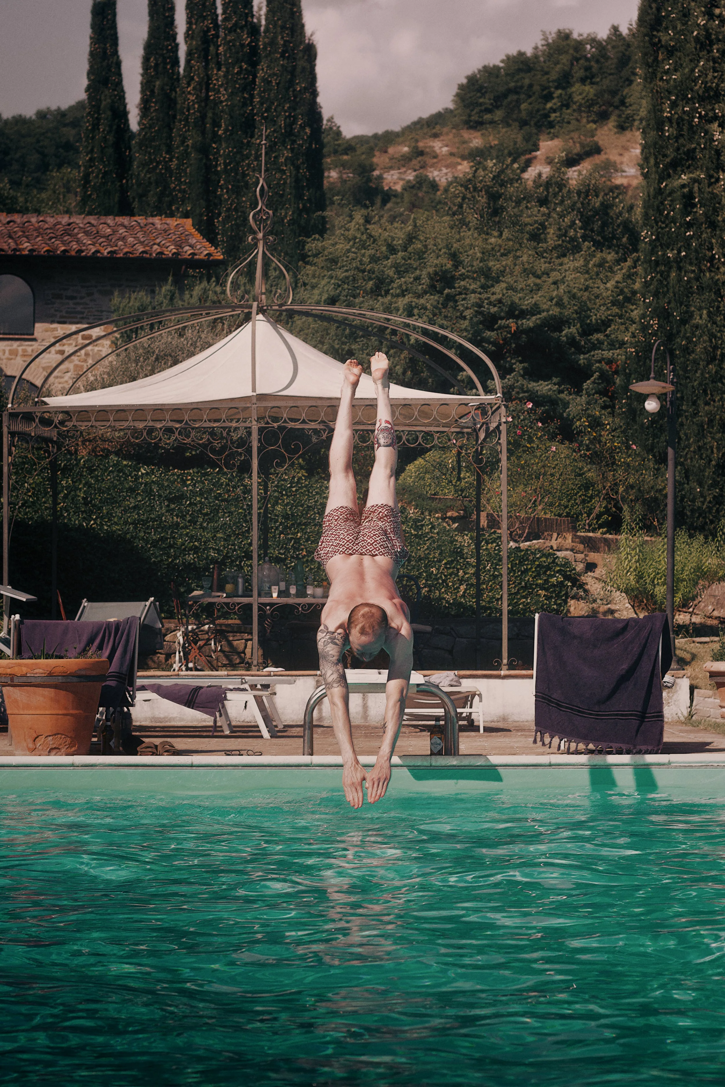 A man performing a handstand at the edge of a swimming pool, with his legs extended upwards and his head and hands touching the water. In the background, there is a gazebo, trees, and a hillside.
