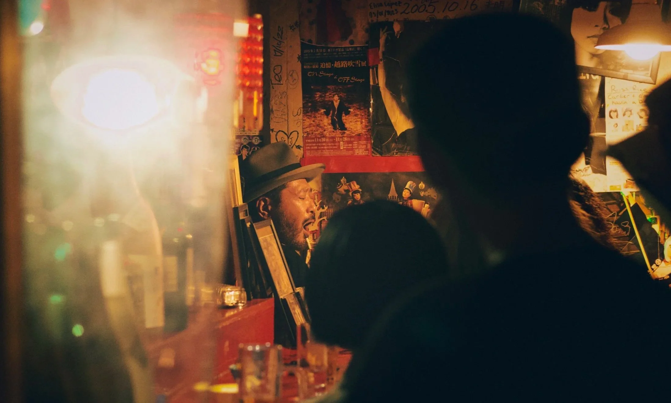 A man with a beard and a hat is singing or speaking in a dimly lit room decorated with posters and graffiti. There is a person with curly hair facing away from the camera on the right, and the scene has a warm, orange glow with a blurred background.