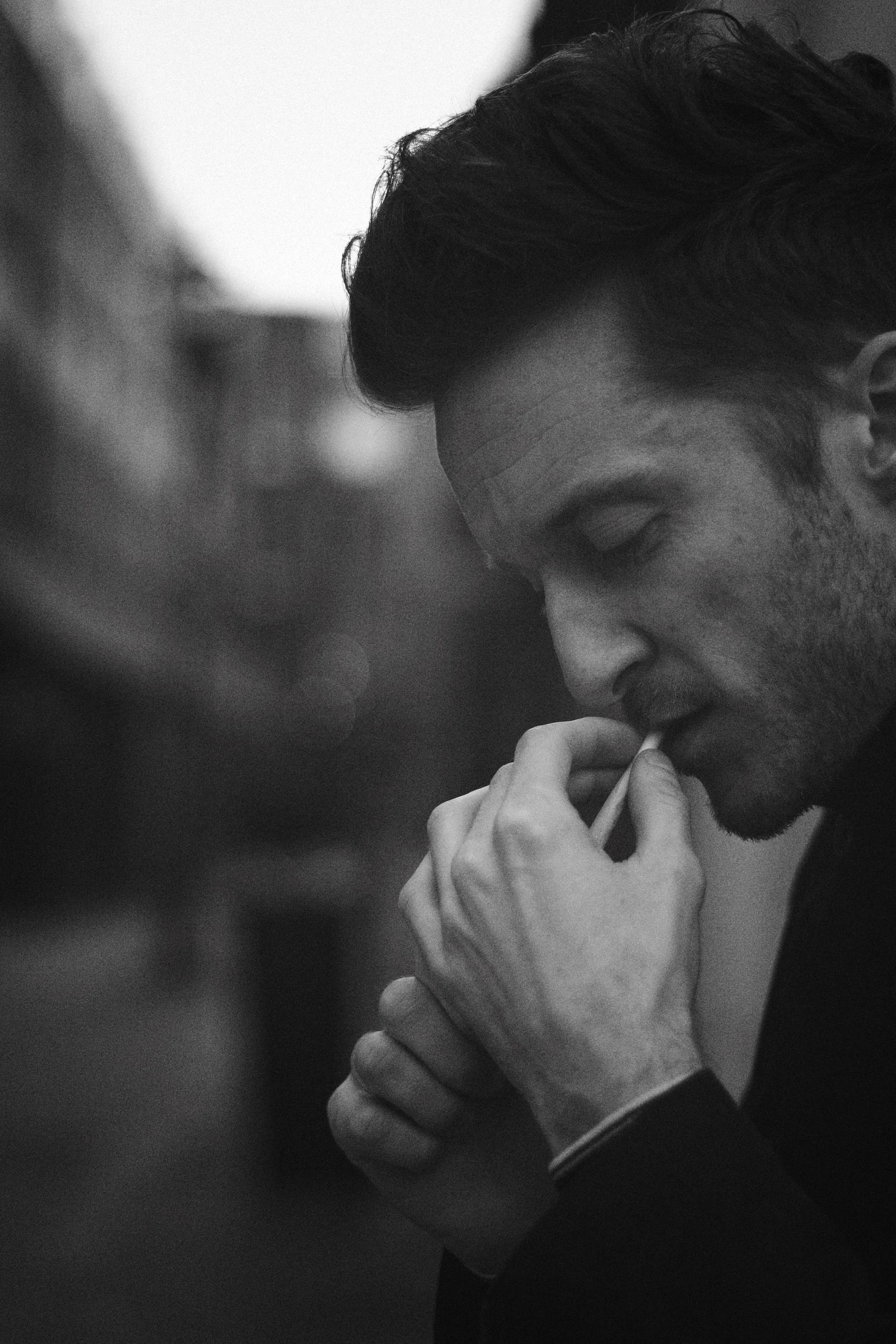 A black and white photo of a man with dark hair, eyes closed, holding a cigarette near his lips, with his hand gripping the cigarette and his other hand holding his chin, in a contemplative pose.