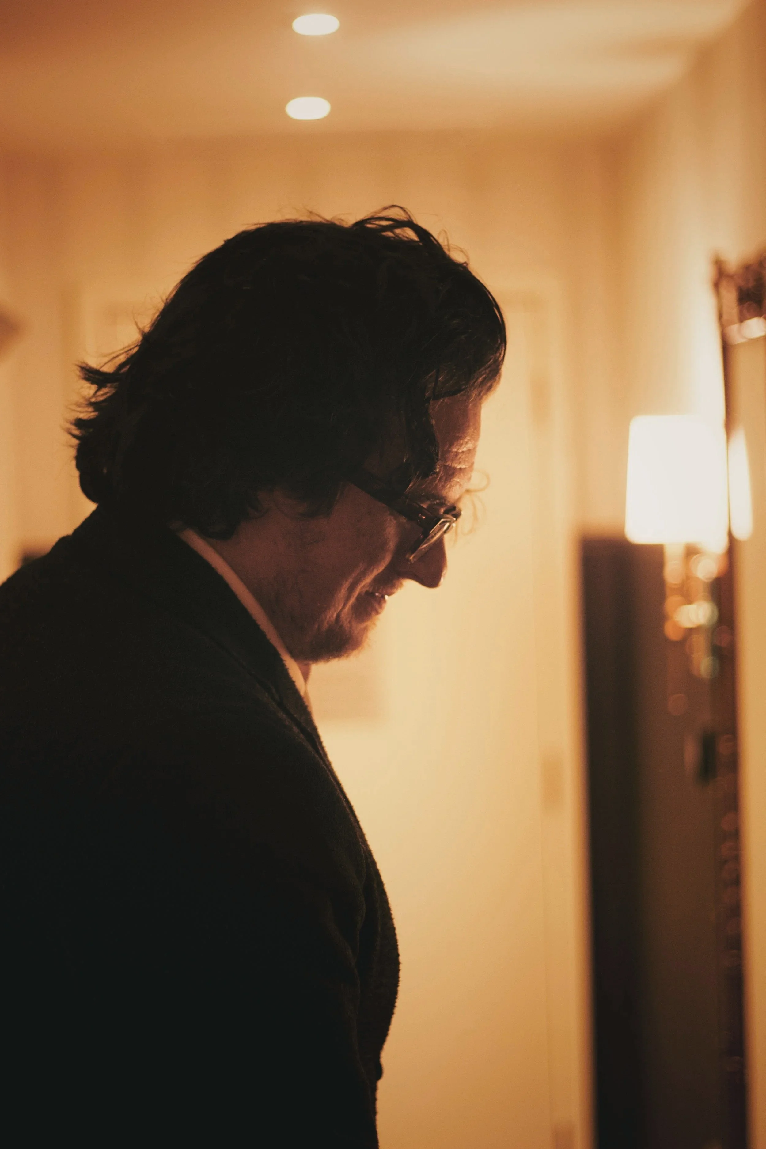 Profile of a man with dark, curly hair and glasses, standing indoors in warm lighting, smiling and looking downward.