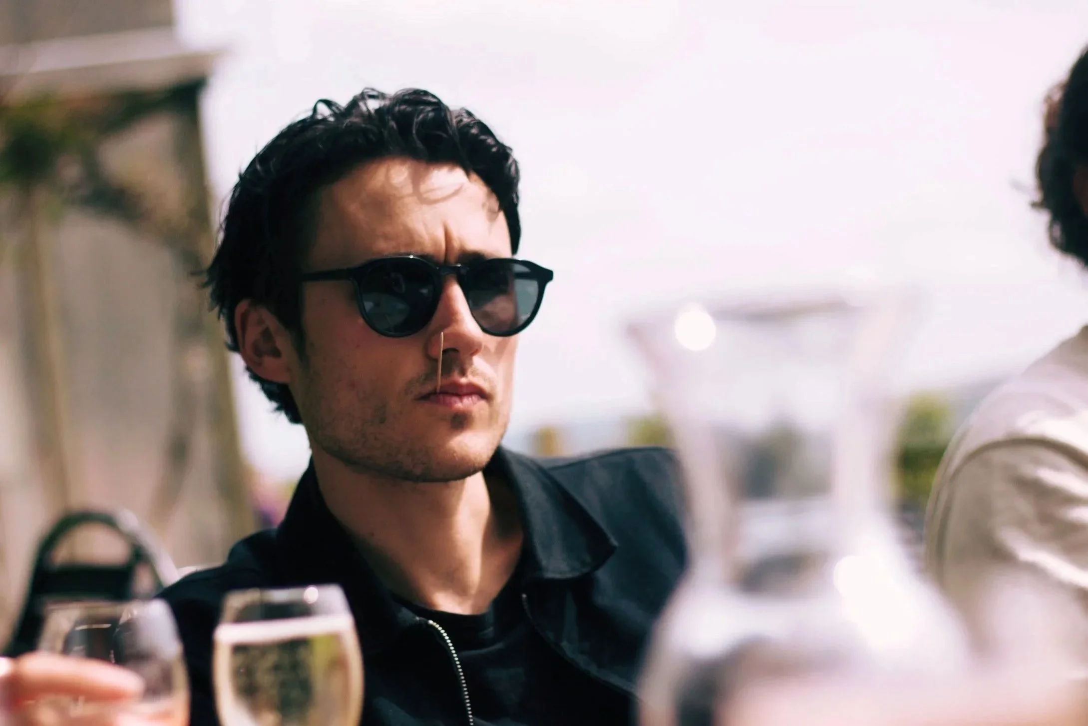 A man with dark, curly hair and wearing black sunglasses, dressed in a black jacket, is holding a glass in his hand, possibly at an outdoor gathering or party during daytime.