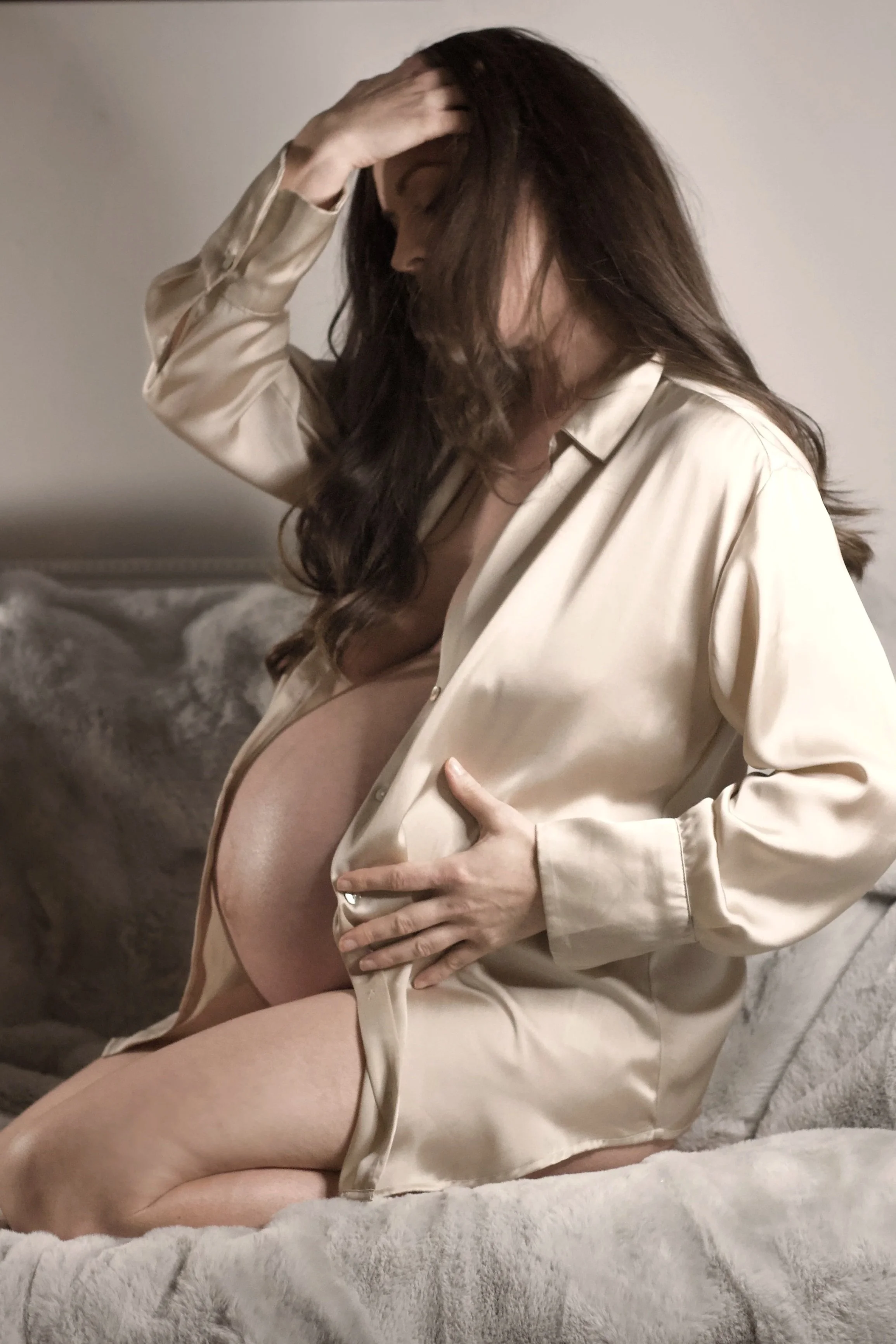 A woman with dark hair, wearing a satin beige robe, is sitting on a plush, light-colored blanket with her hand on her forehead and her other hand resting on her pregnant belly, in a softly lit room.