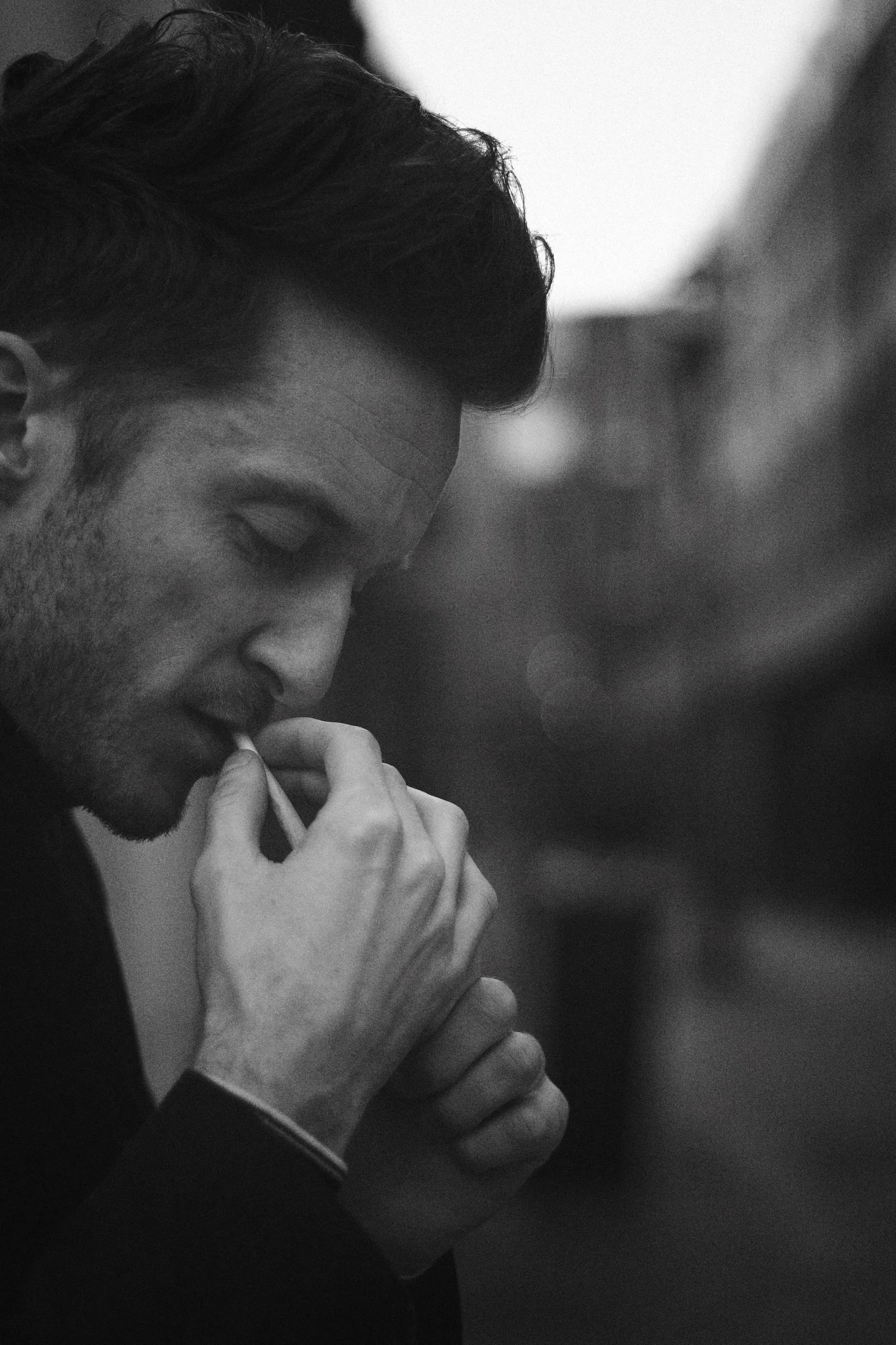 A black and white photo of a man with dark hair, eyes closed, holding a cigarette near his lips, with his hand gripping the cigarette and his other hand holding his chin, in a contemplative pose.