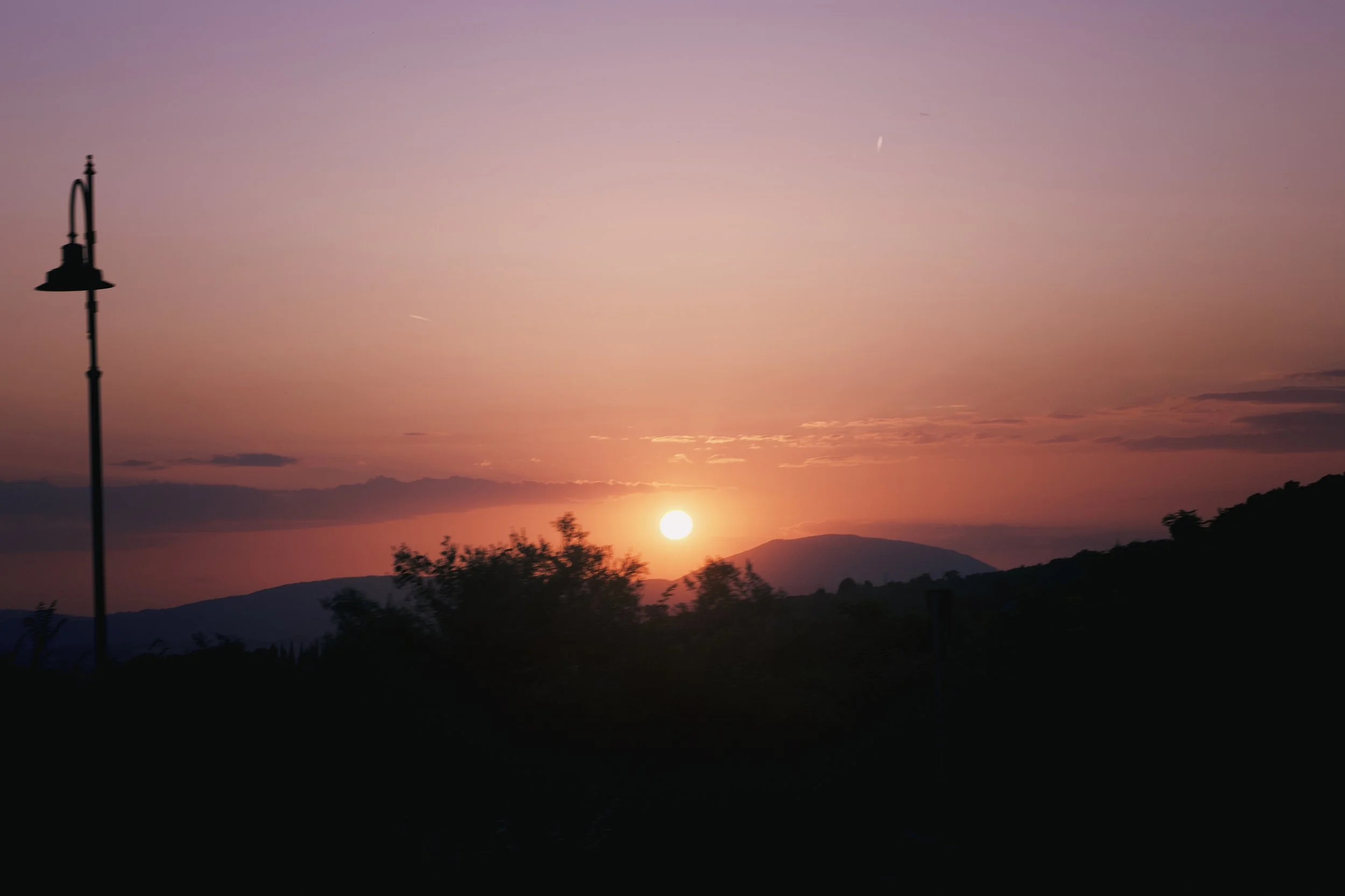 Sunset over mountains with a pink sky, a streetlamp on the left, silhouettes of trees and hills in the foreground.