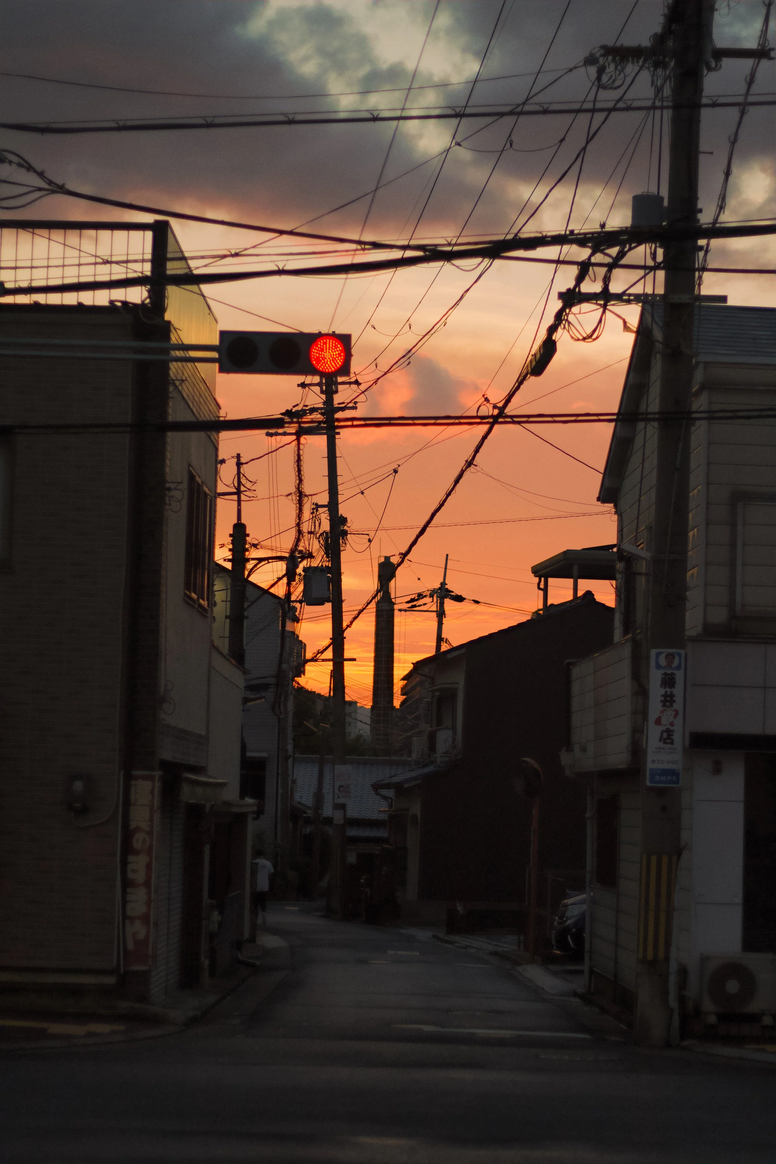 A street scene at sunset with an overcast sky, showing utility poles, wires, and a traffic light displaying red. Buildings line the street, with some signs and windows visible.