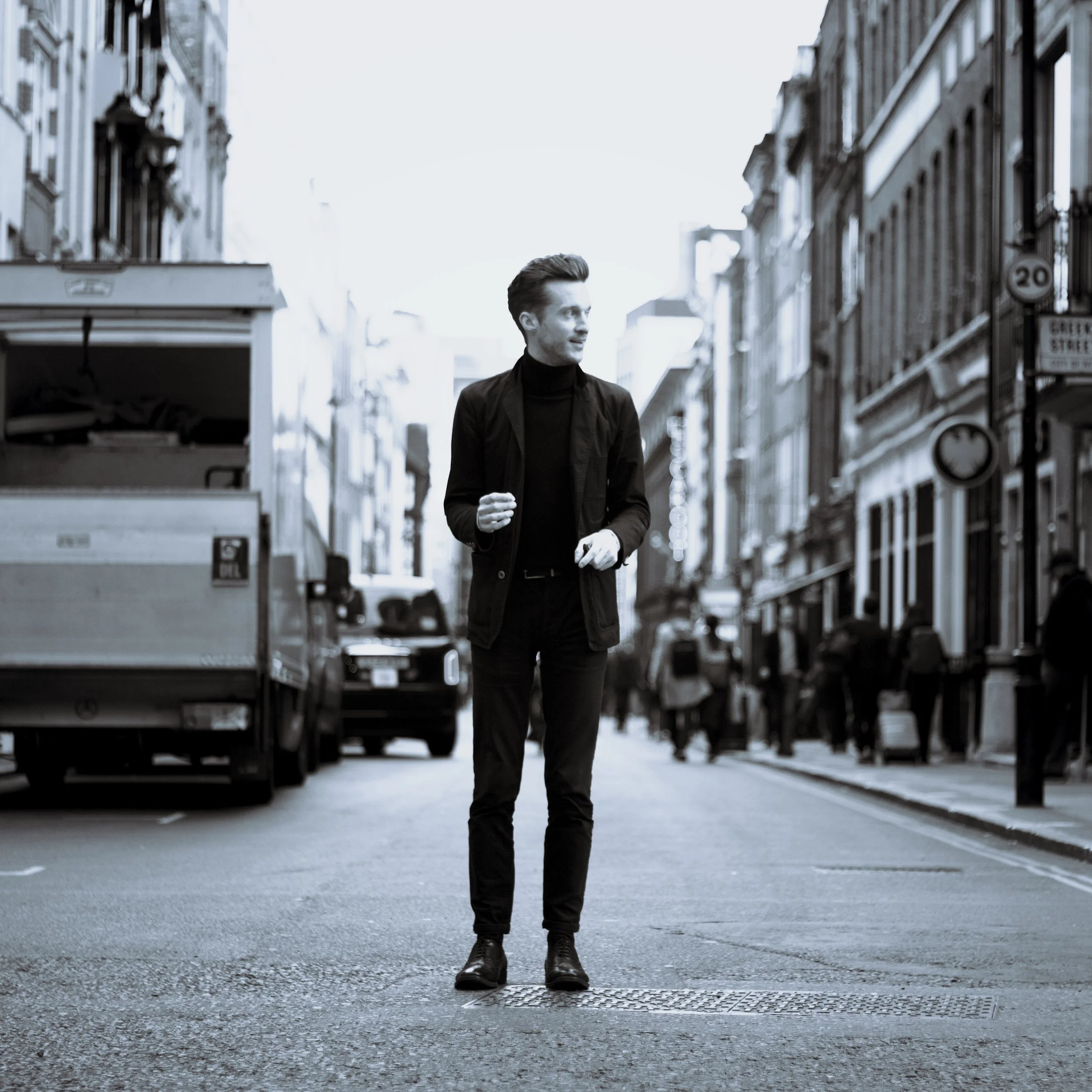A young man with styled hair and wearing a black turtleneck and jacket walking outdoors in an urban setting with buildings and parked cars in the background.