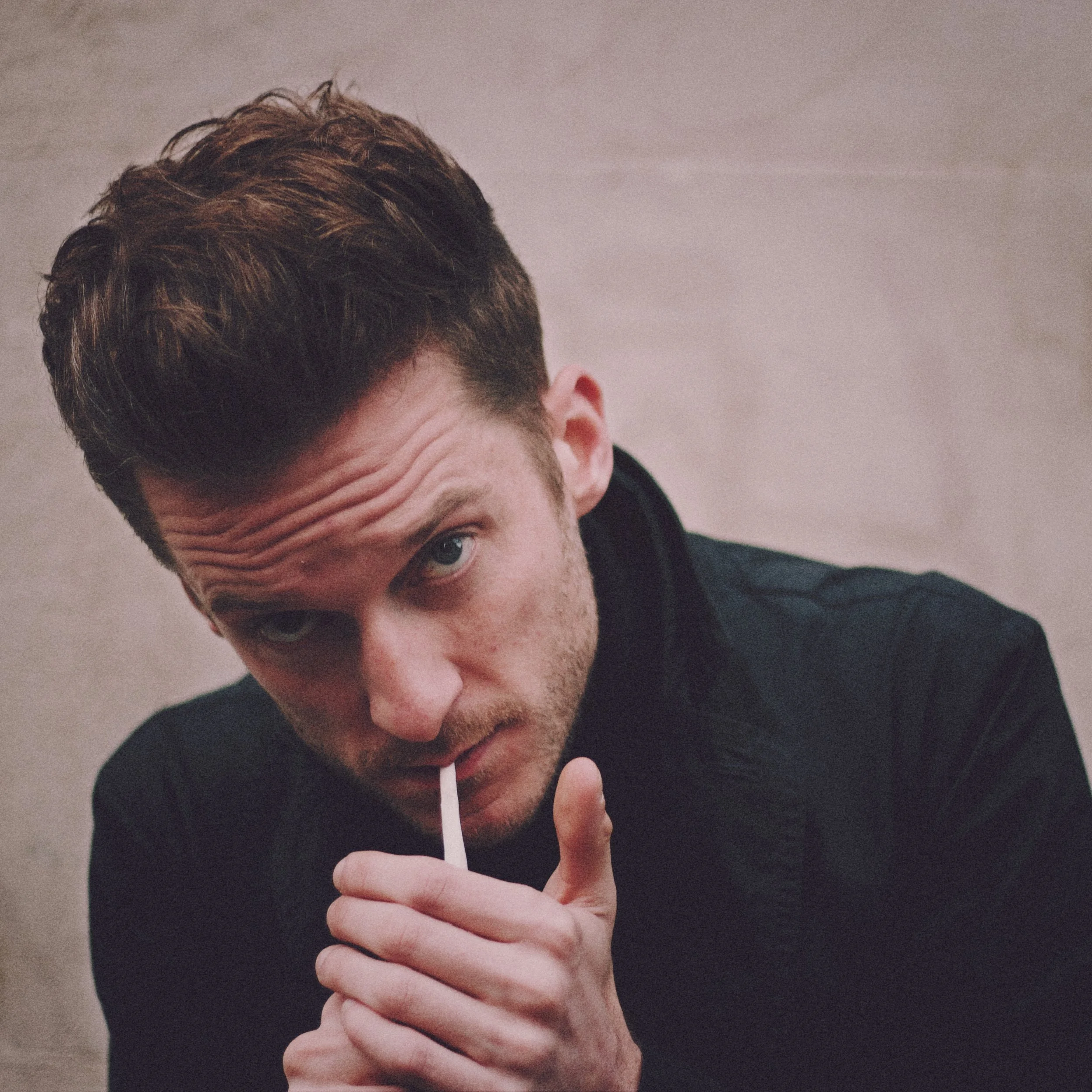 Close-up of a man with brown hair and a beard, holding a cigarette near his lips with his hand, wearing a dark jacket.