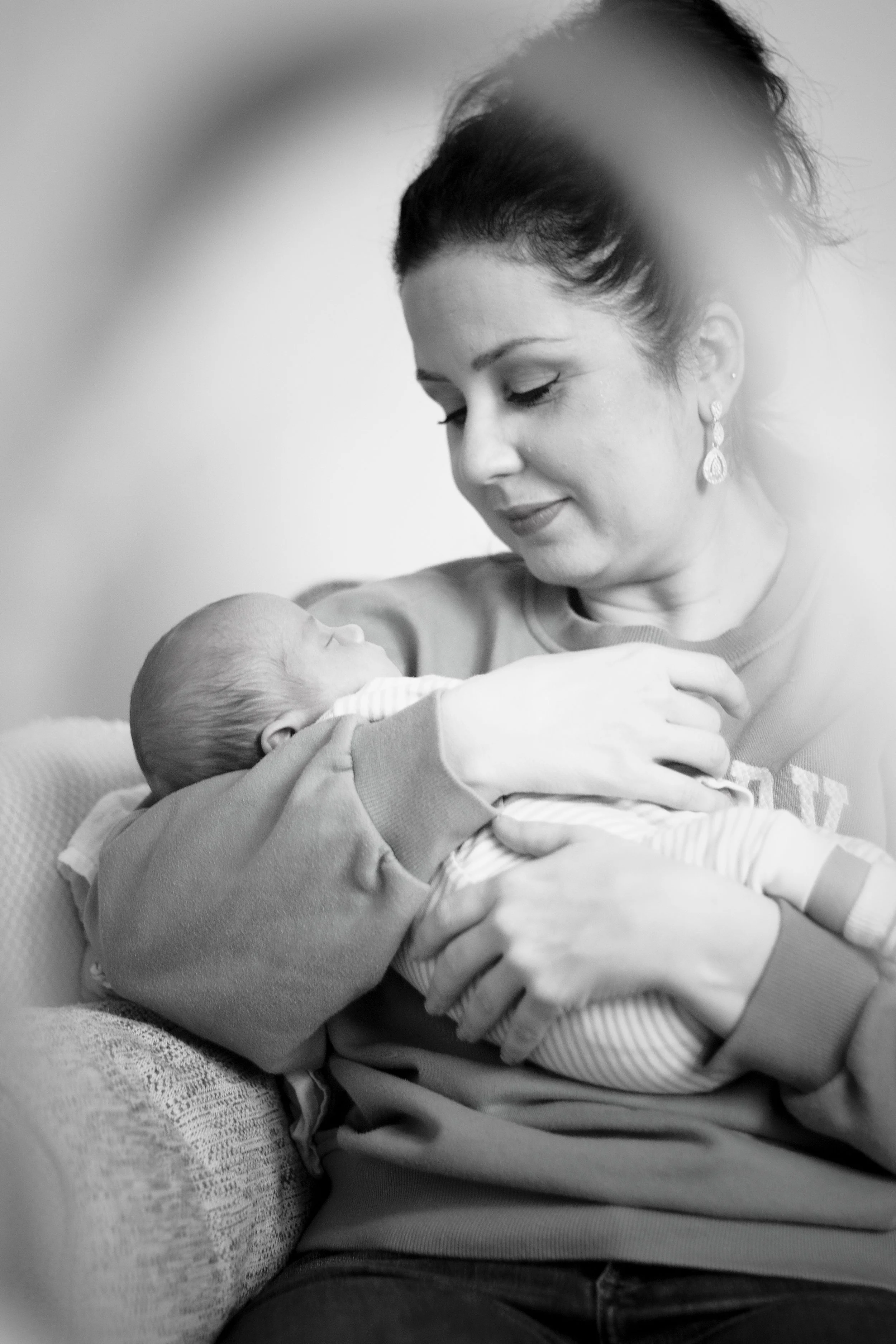 Black and white photograph of a woman holding a newborn baby, looking down tenderly at the baby, who is wrapped in a striped blanket. The woman has her eyes closed and is wearing elegant earrings.