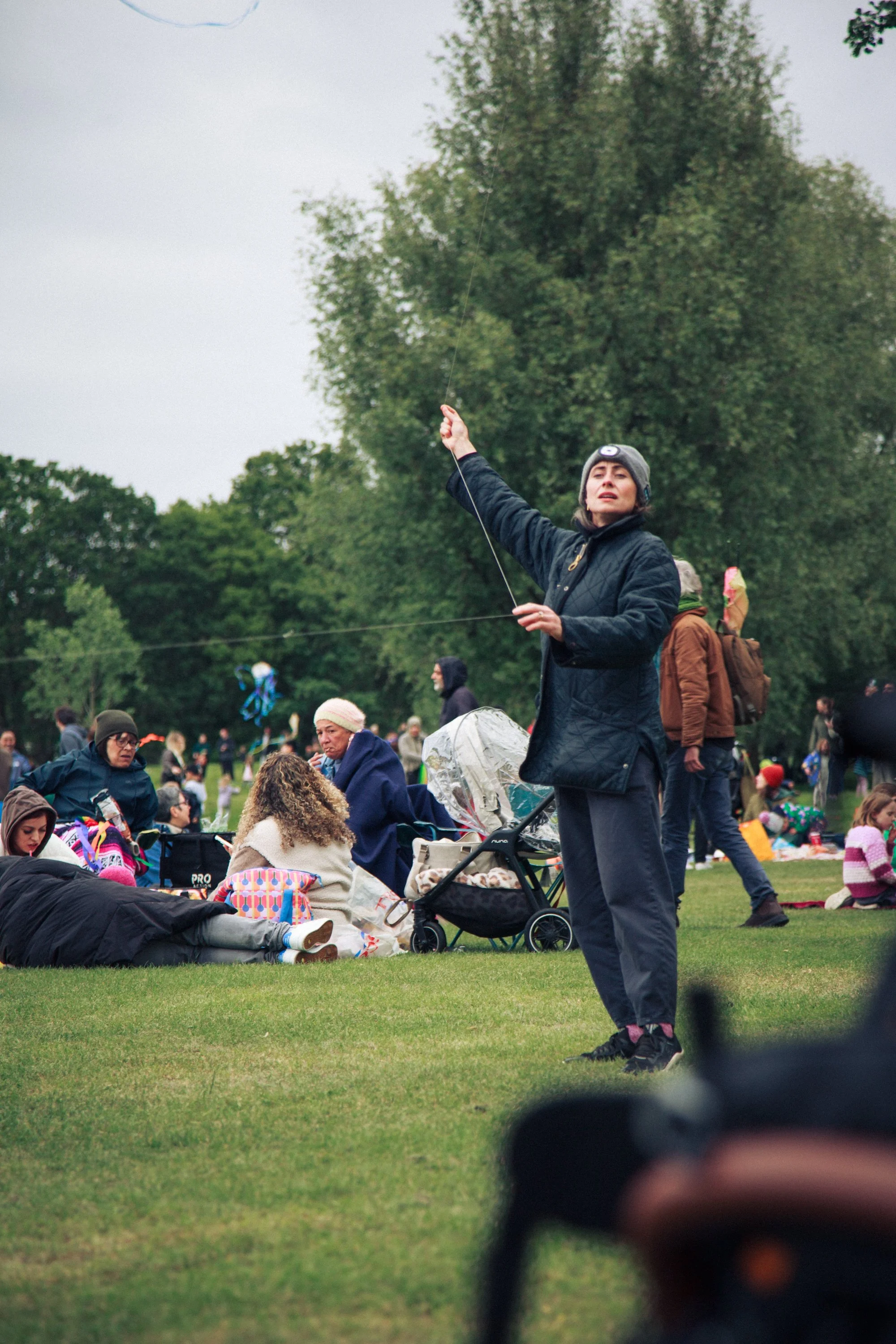 A group of people gathered outdoors in a park, with some sitting on the grass and others standing. A woman in the center is holding a kite string aloft, and there are trees in the background.