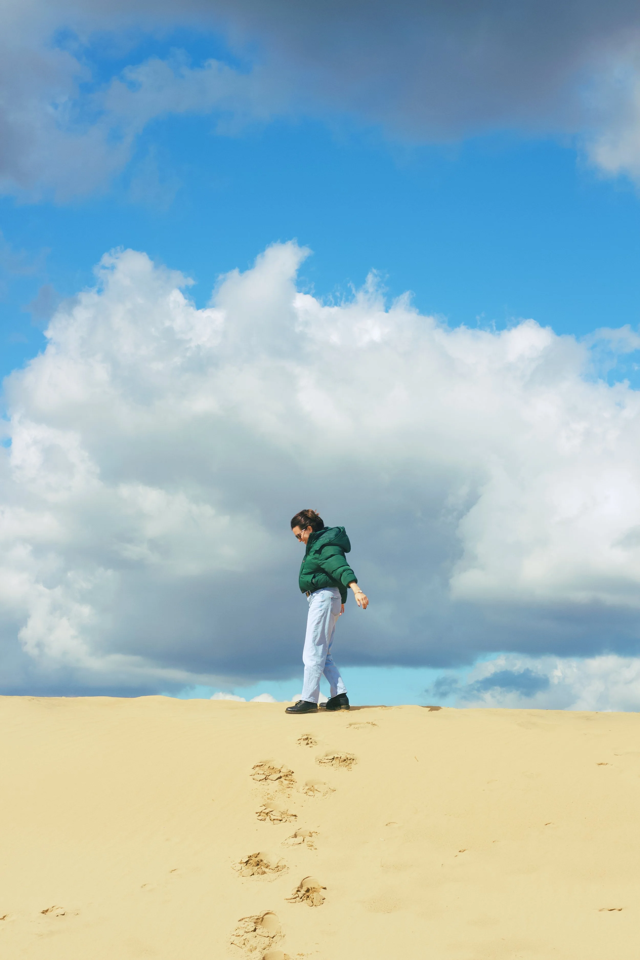 A person in a green jacket and light blue jeans walking on sandy terrain with footprints, under a partly cloudy sky.