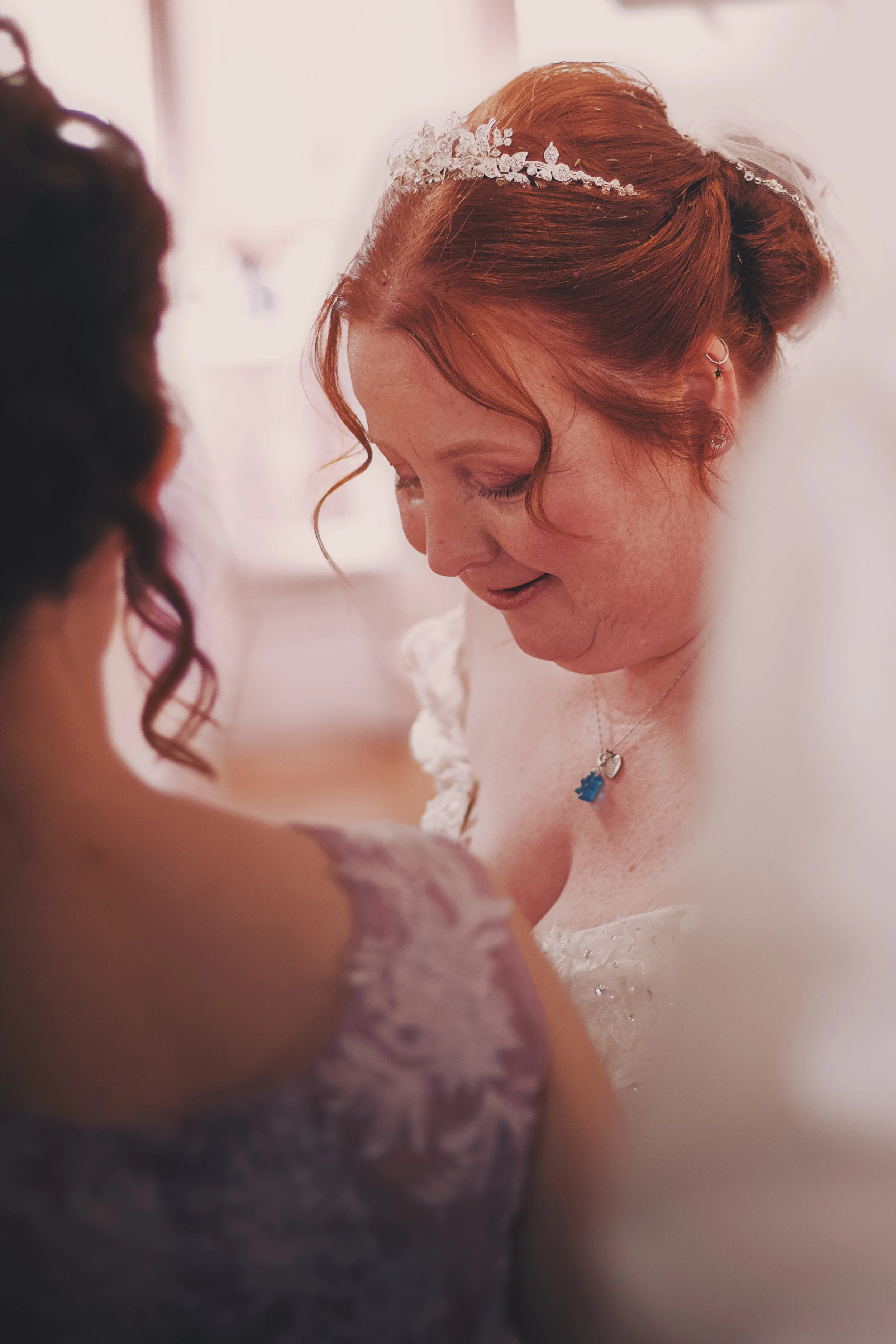 A bride with red hair wearing a tiara, earrings, and a necklace, smiling and looking down, with a young girl with dark, curly hair in the foreground.