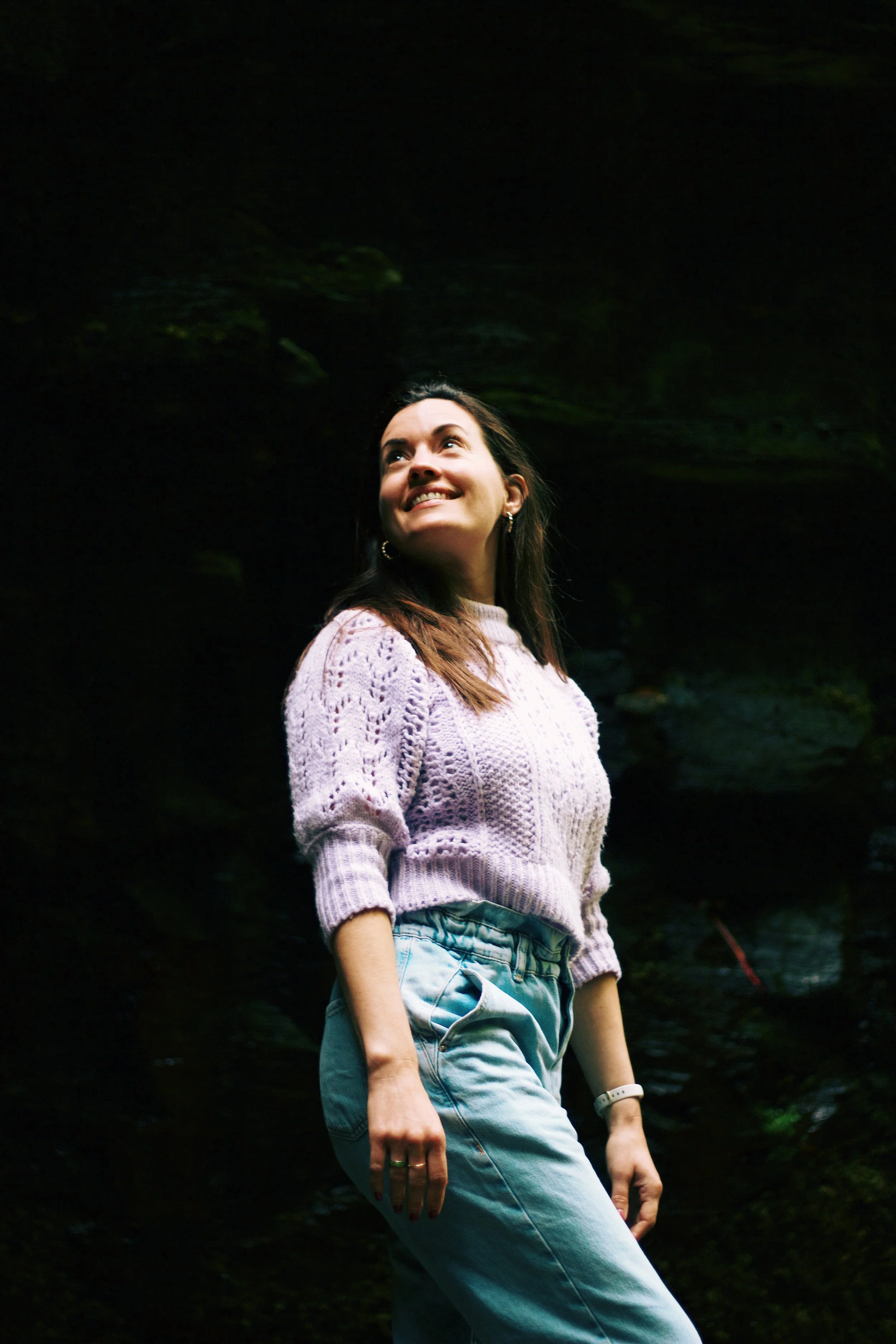 A woman smiling and looking up, wearing a light pink knitted sweater, light blue jeans, and a white watch, standing outdoors with dark green foliage in the background.