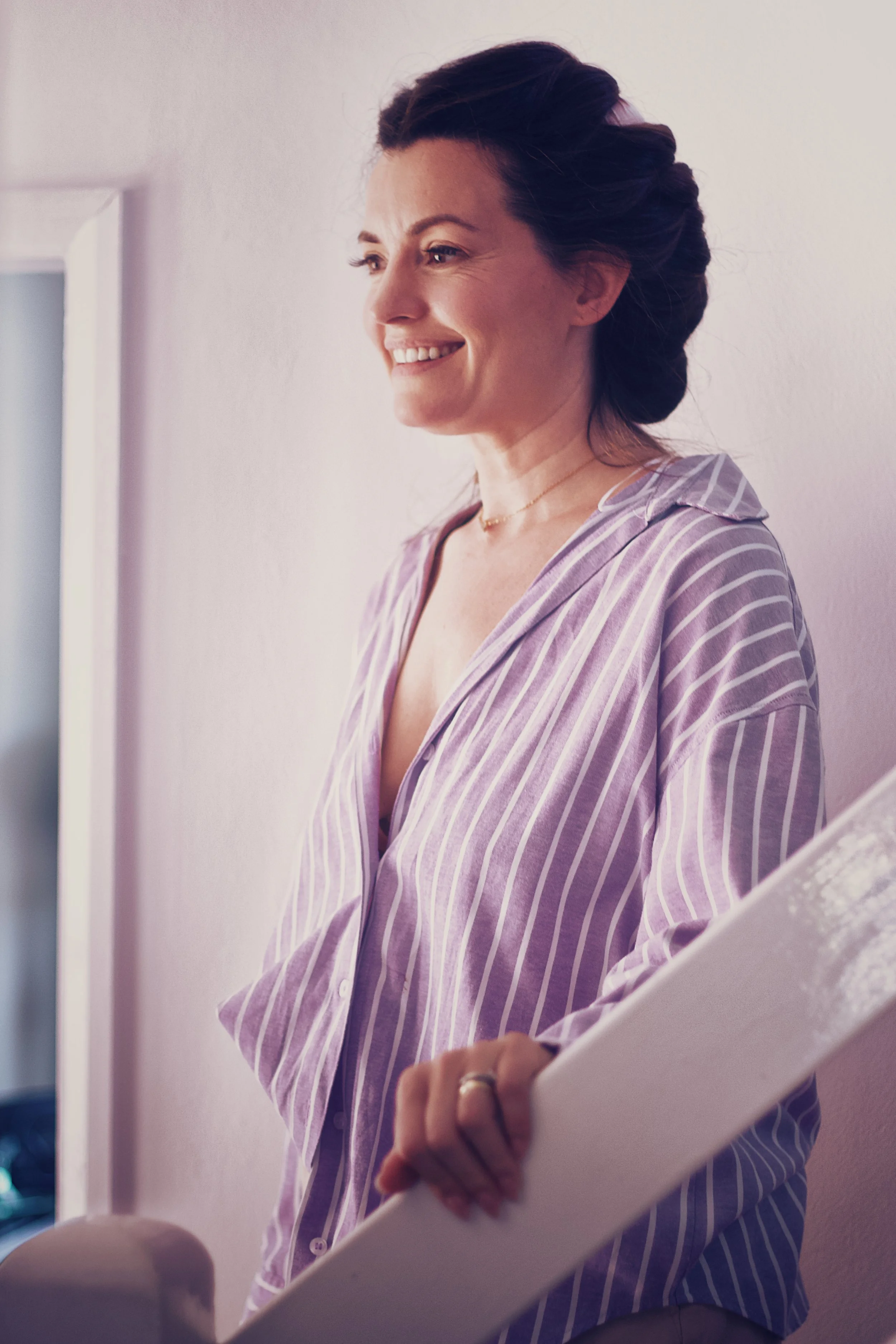 A woman with dark hair styled in an updo, wearing a purple and white striped pajama top, smiling and looking at a mirror while holding a white tablet.