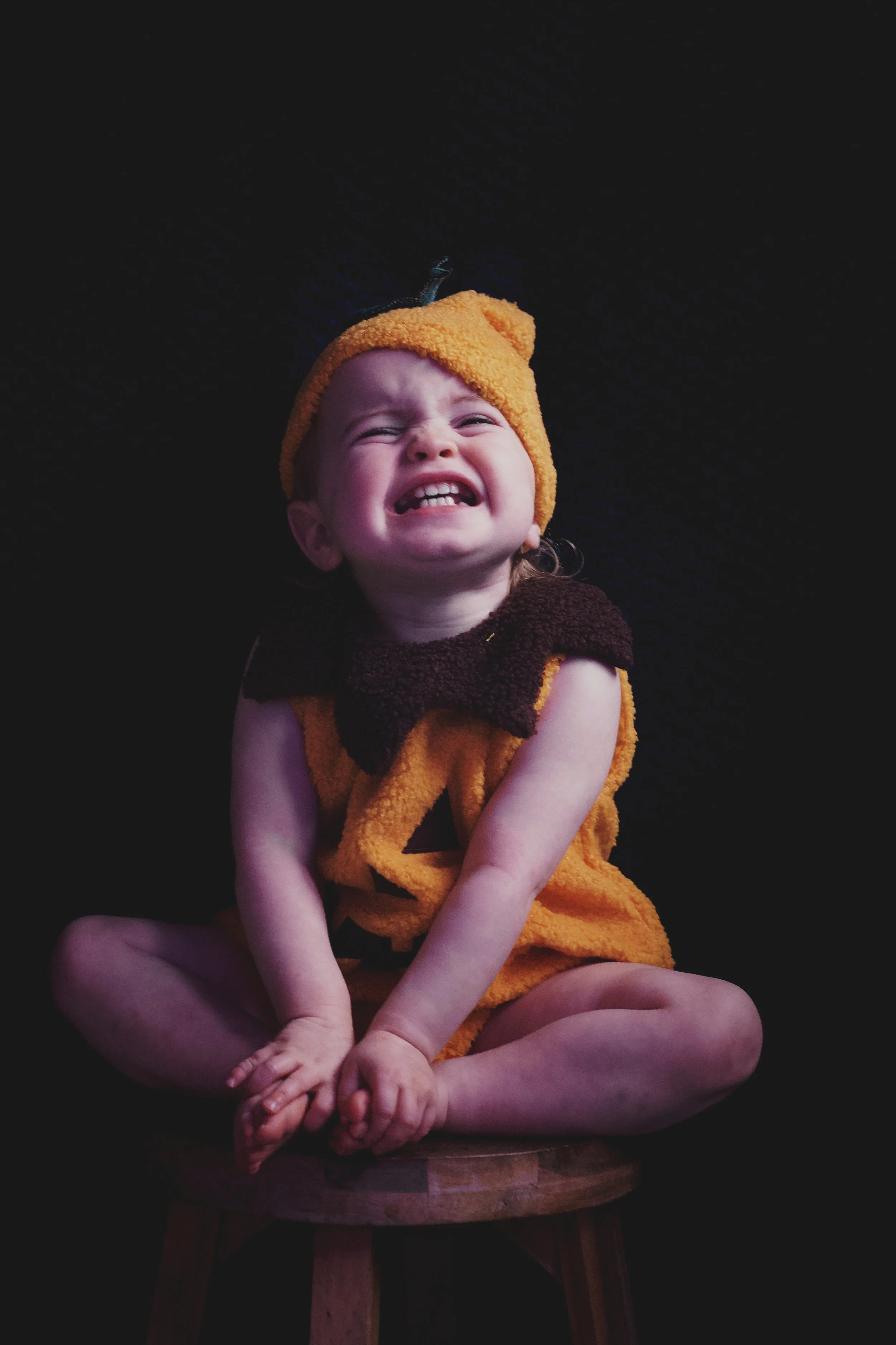 A young child in a pumpkin costume, making a silly face with eyes squinted and mouth open, sitting on a wooden seat with a black background.