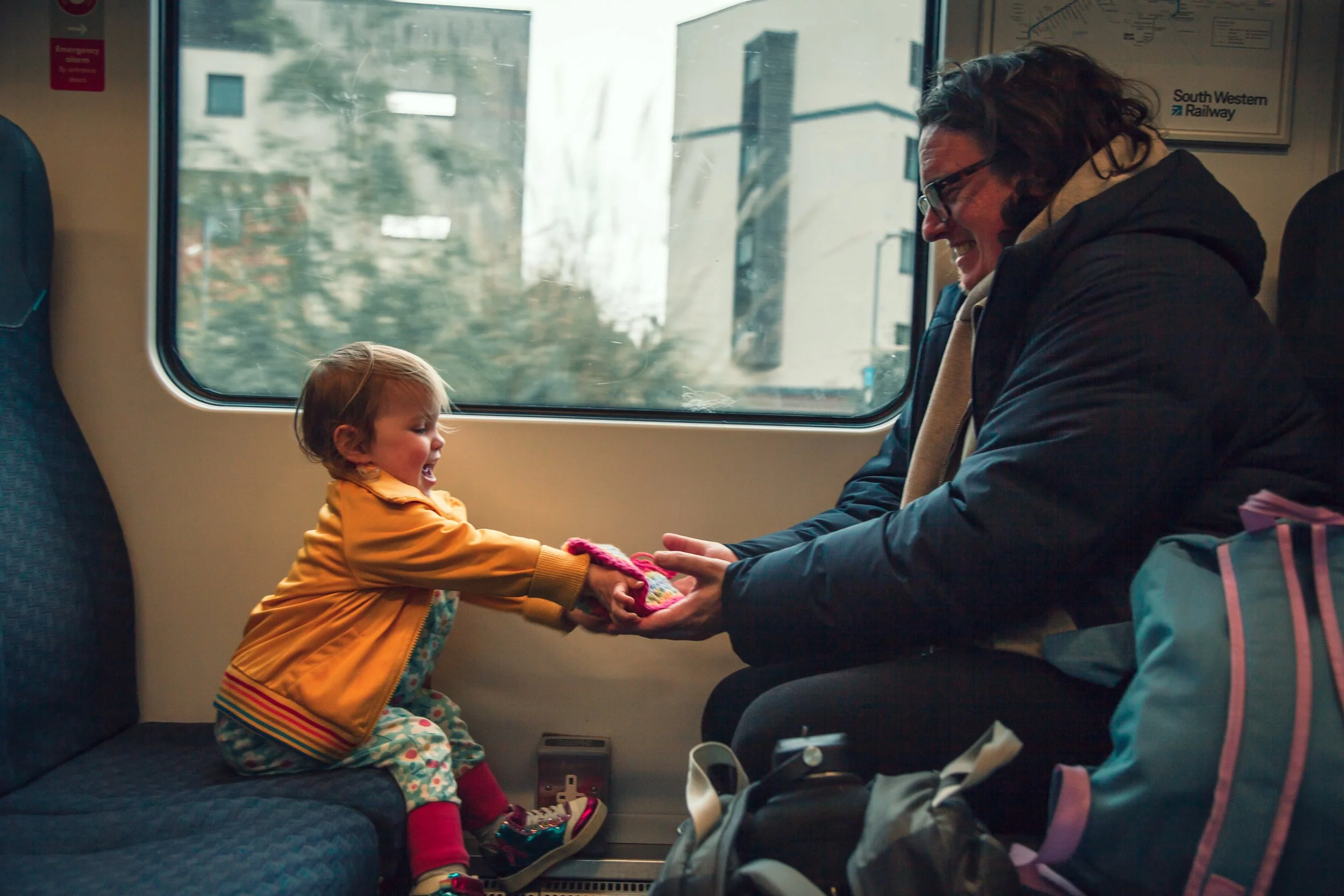 A child and an adult on a train. The child, wearing a yellow jacket and colorful pants, is smiling and holding the adult's hands. The adult, wearing a dark jacket and glasses, is smiling and looking at the child. They are sitting by a window with a c