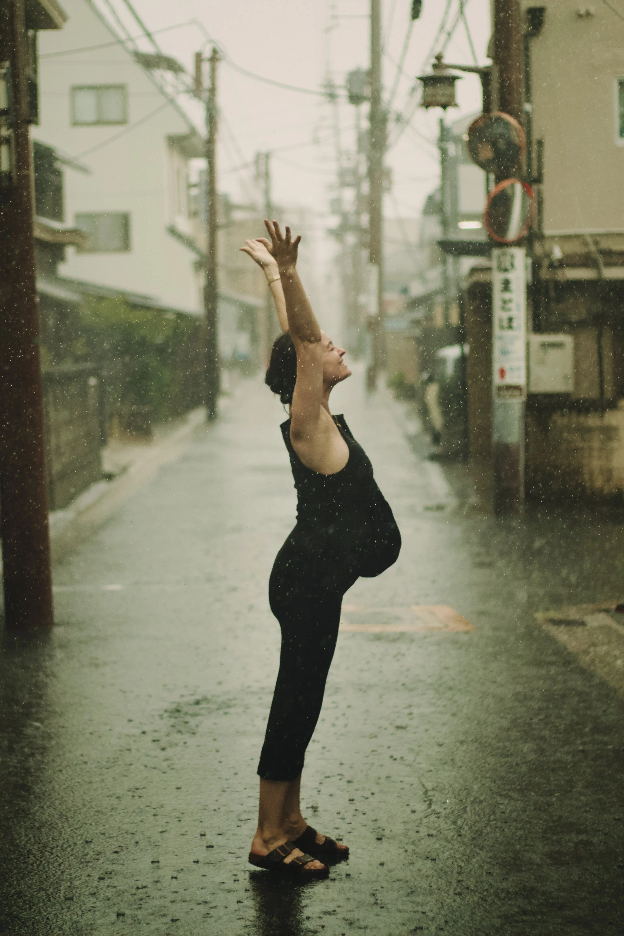 A woman standing on a street during a rainstorm, with her arms raised above her head, smiling and looking upward. She is wearing a black dress and sandals, with the rain pouring all around her.