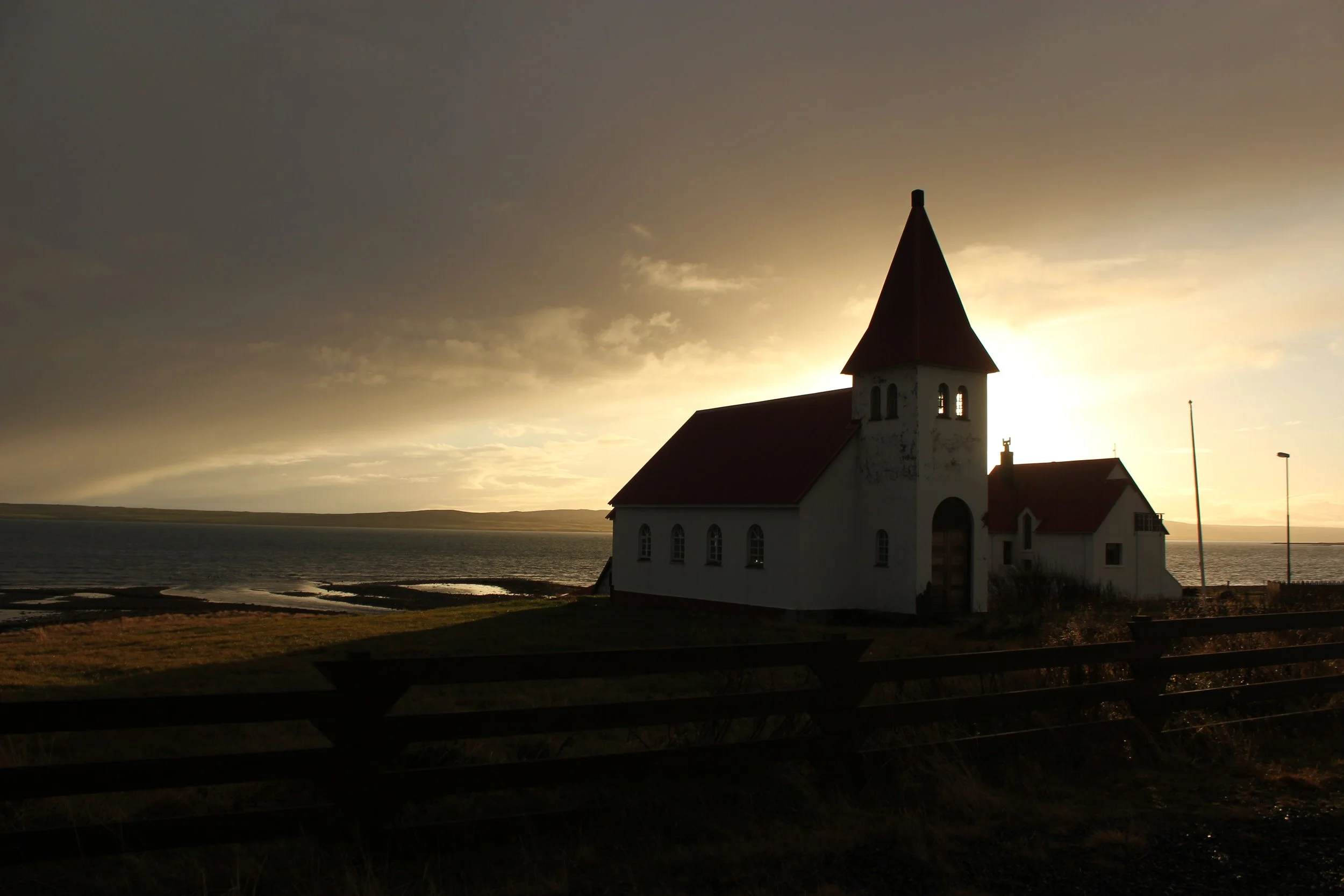 Church with a tall steeple by the water during sunset, with a cloudy sky and a distant shoreline in the background.
