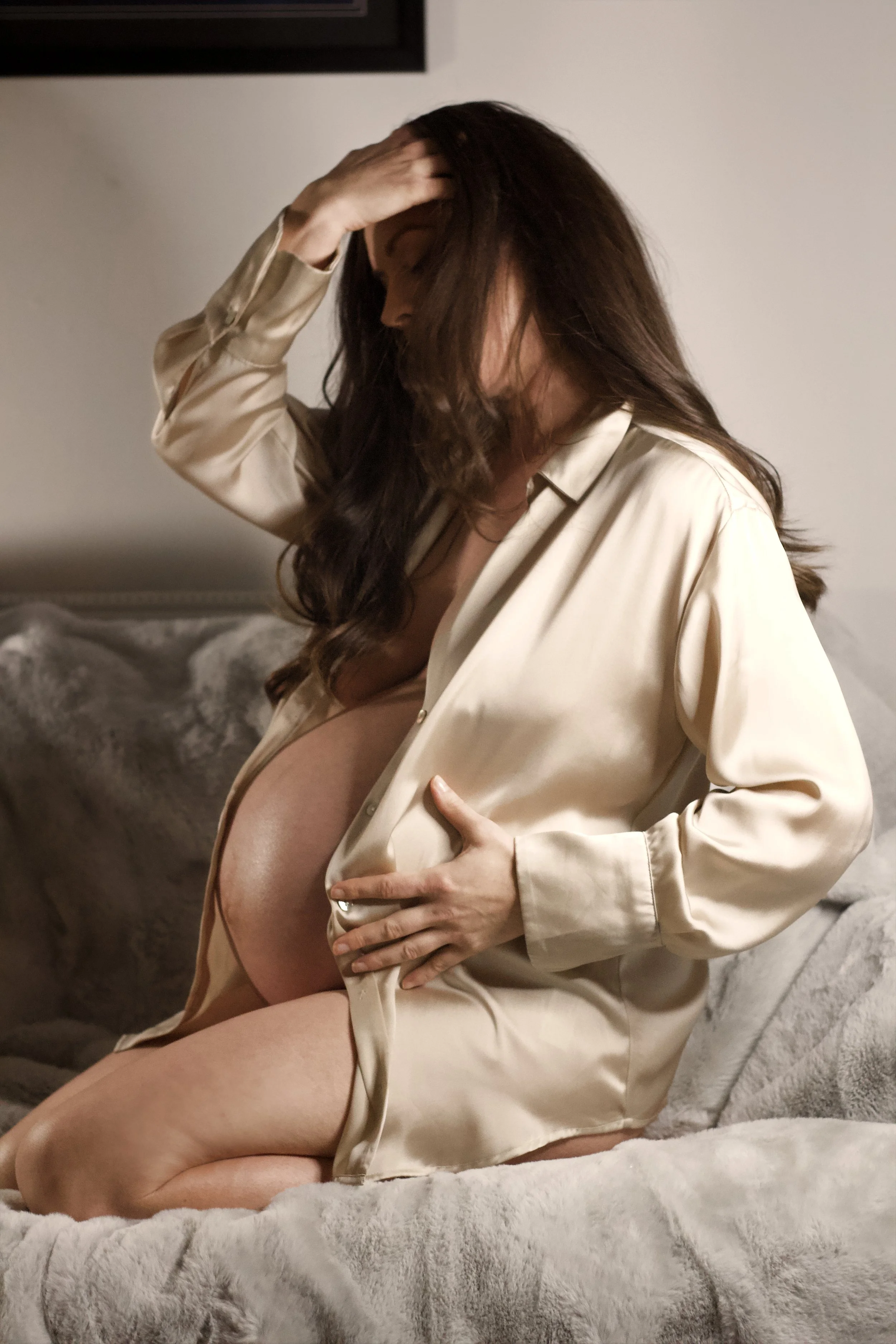 A woman with dark hair, wearing a satin beige robe, is sitting on a plush, light-colored blanket with her hand on her forehead and her other hand resting on her pregnant belly, in a softly lit room.