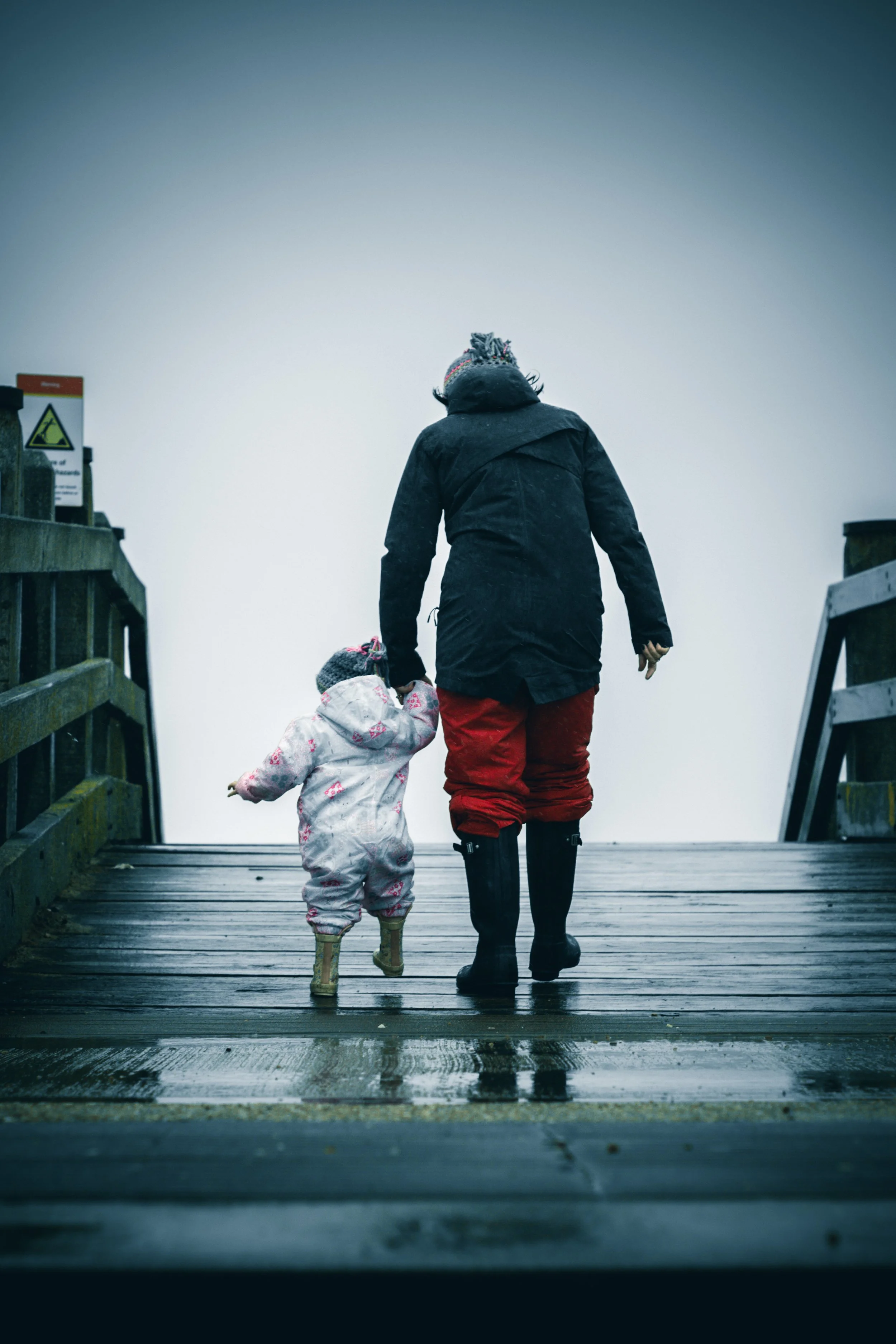 A person and a child walk on a wooden pier or boardwalk with stairs on either side, dressed in waterproof clothing and boots, suggesting rainy or muddy weather, with a foggy or overcast sky.