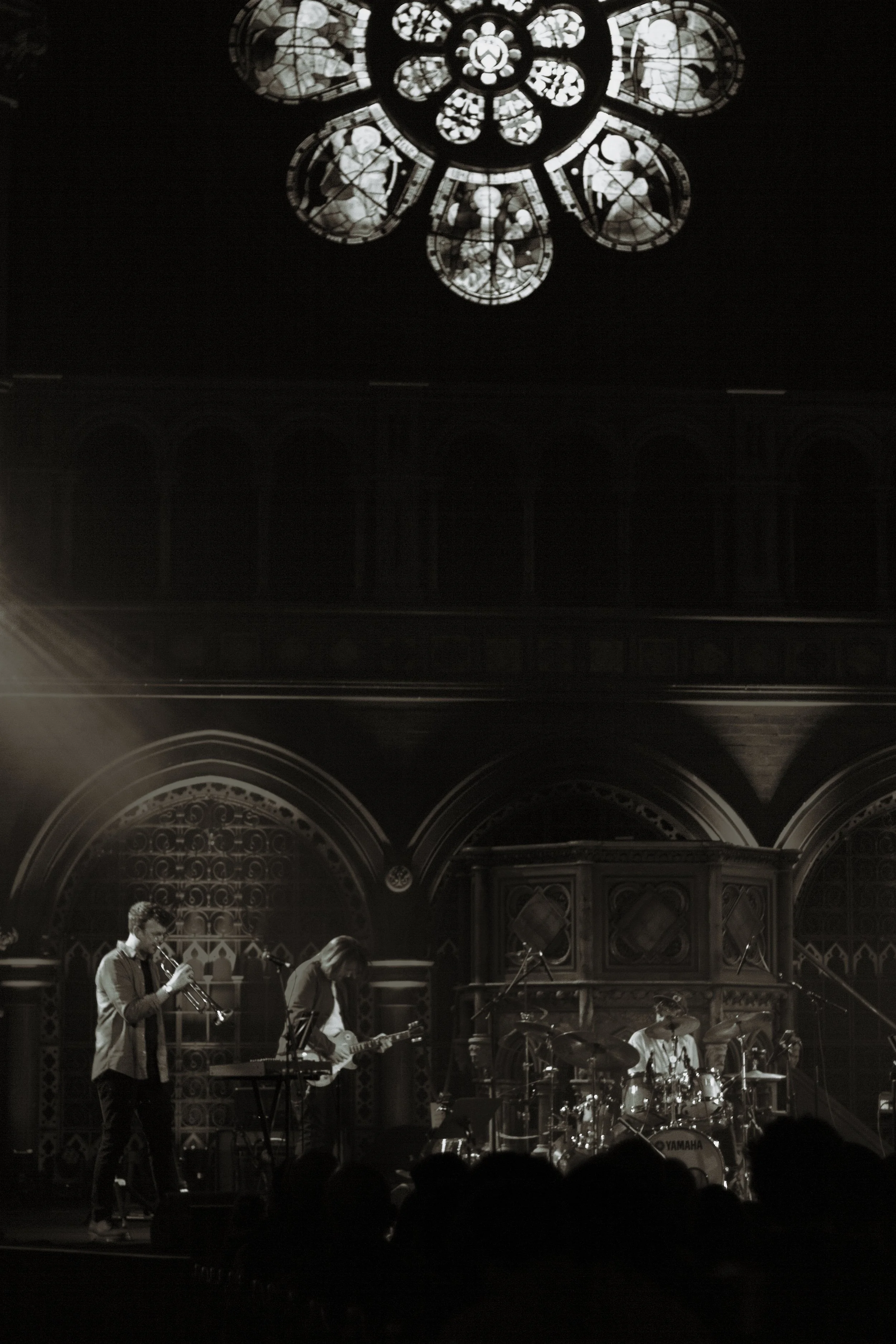 A black and white photo of a live band performing on stage inside a church, with a large stained glass window above them. The band members are playing various instruments, including drums, keyboard, guitar, and trumpet.