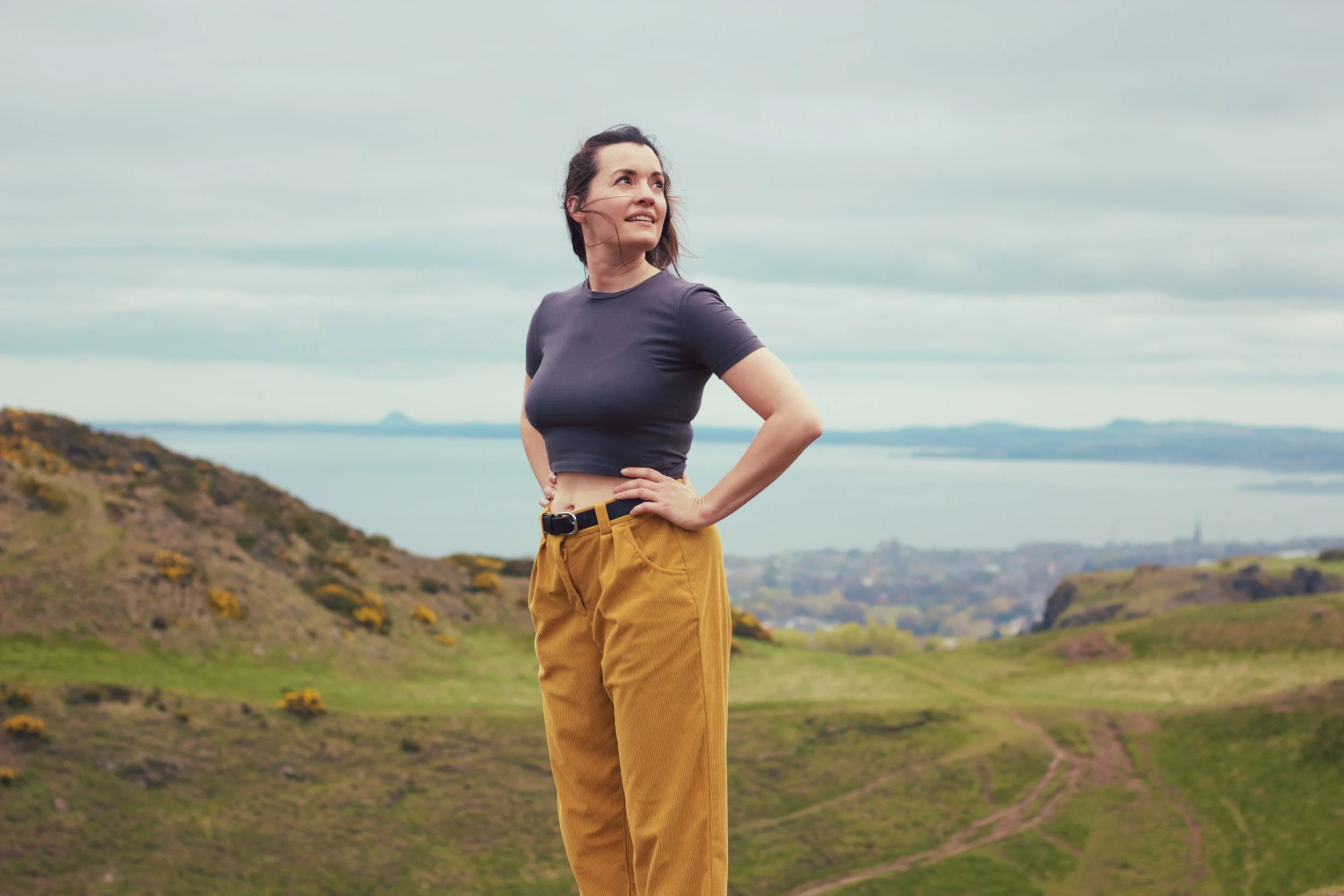 A woman standing outdoors on a hillside, looking into the distance with a slight smile, with a body of water and hilly landscape in the background.