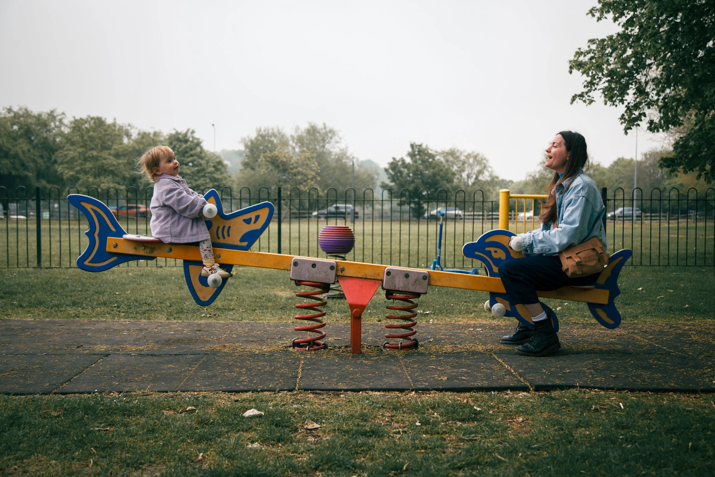A young girl and a woman are sitting on a spring rocking seesaw at a playground, facing each other, with trees and parked cars in the background.