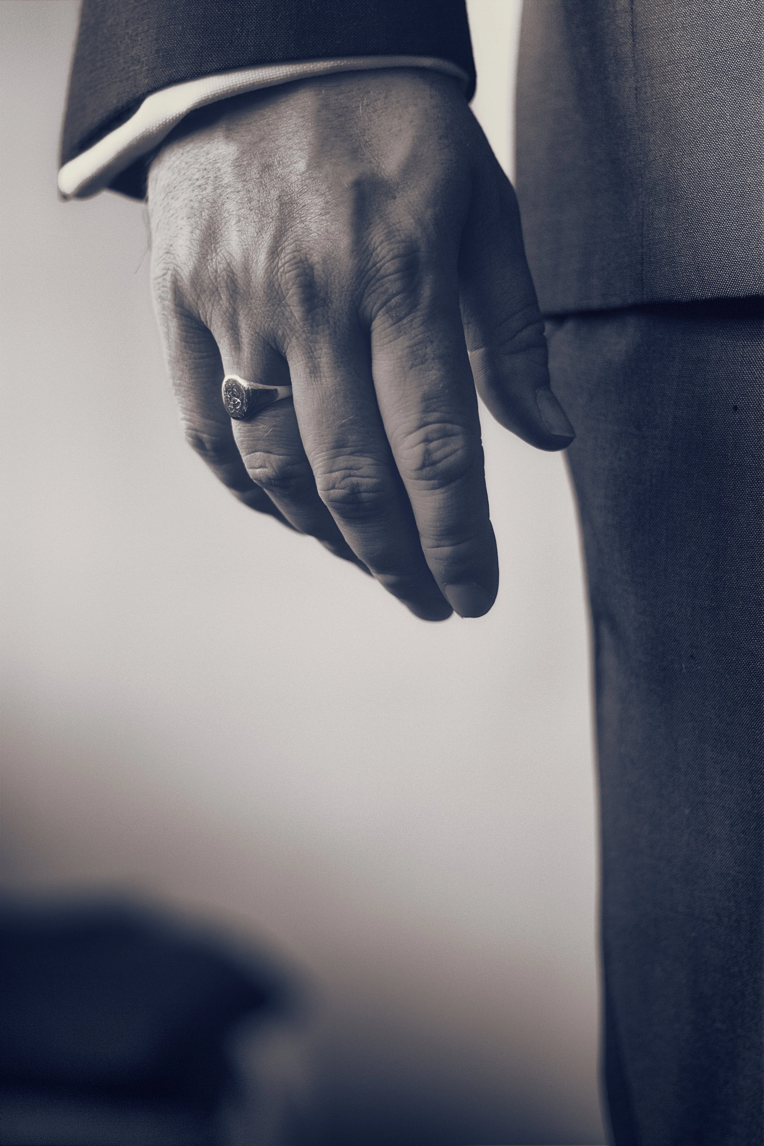 Close-up of a hand with a ring, dressed in a suit, resting against a person's thigh.