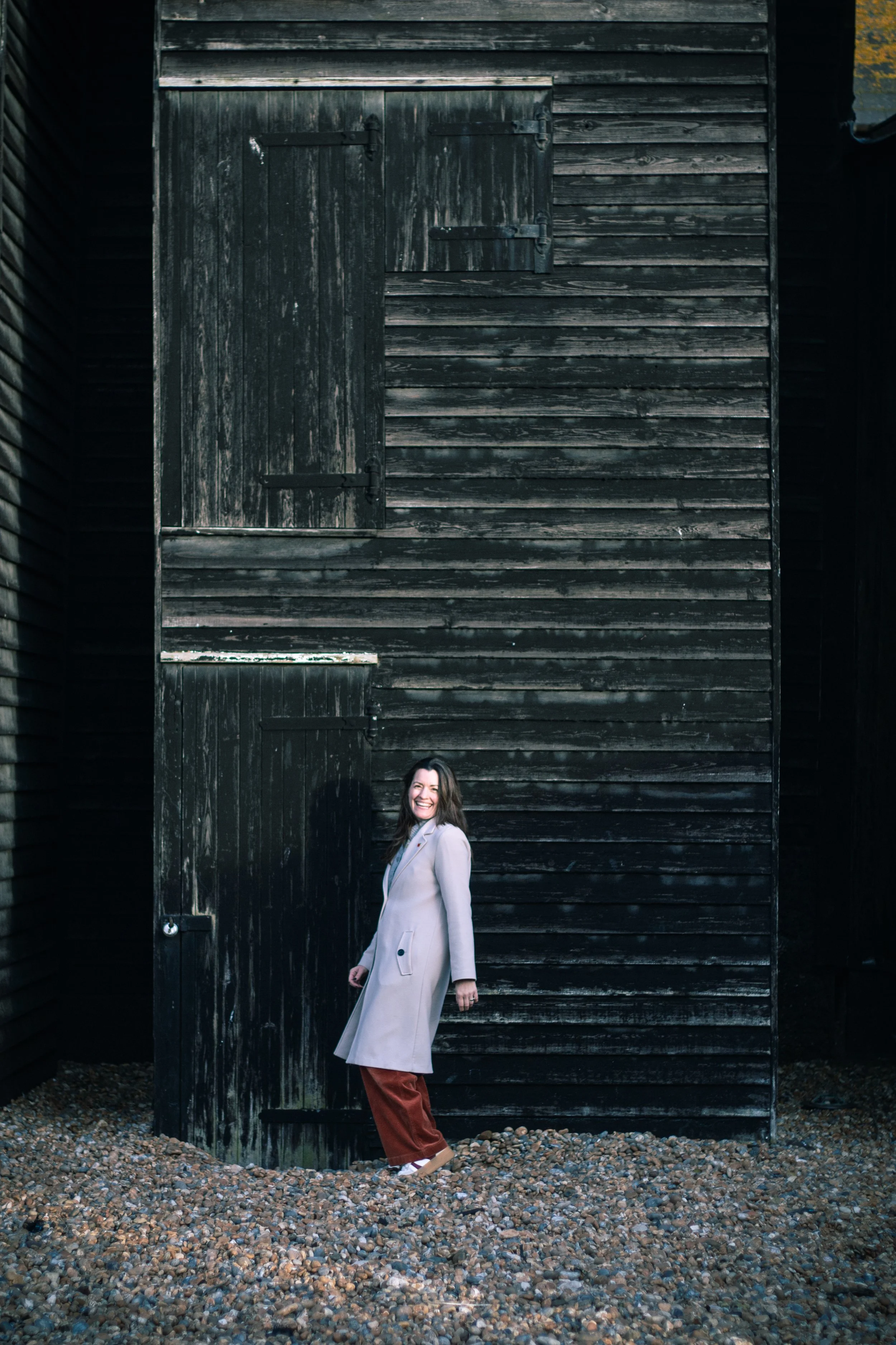 Woman wearing a beige coat and maroon pants smiling and standing outdoors in front of a large wooden dark brown wall.