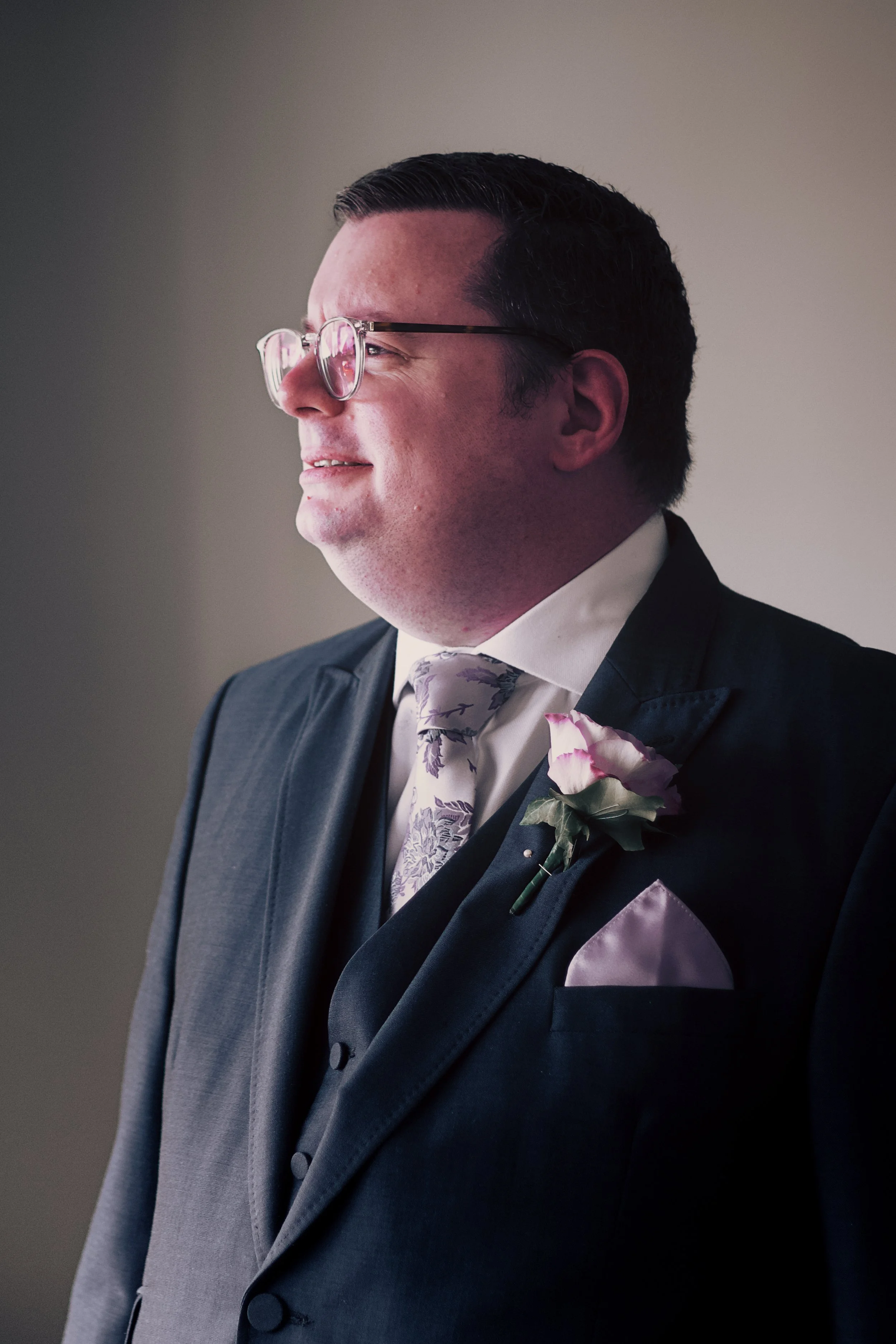 Side profile of a man dressed in a dark suit with a tie, wearing glasses, with a pink flower boutonniere on his lapel, smiling softly against a neutral background.