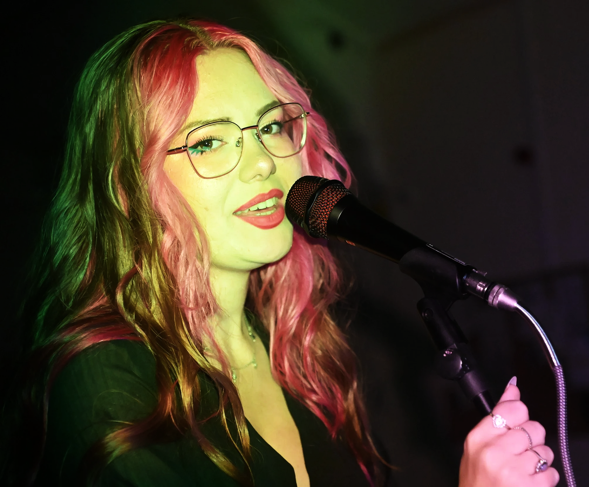 Tay, a female vocalist with long wavy pink and brown hair and glasses - performing live and singing into a microphone under bright green stage lights. She is wearing a black v-neck dress and looks at the camera whilst singing into a microphone