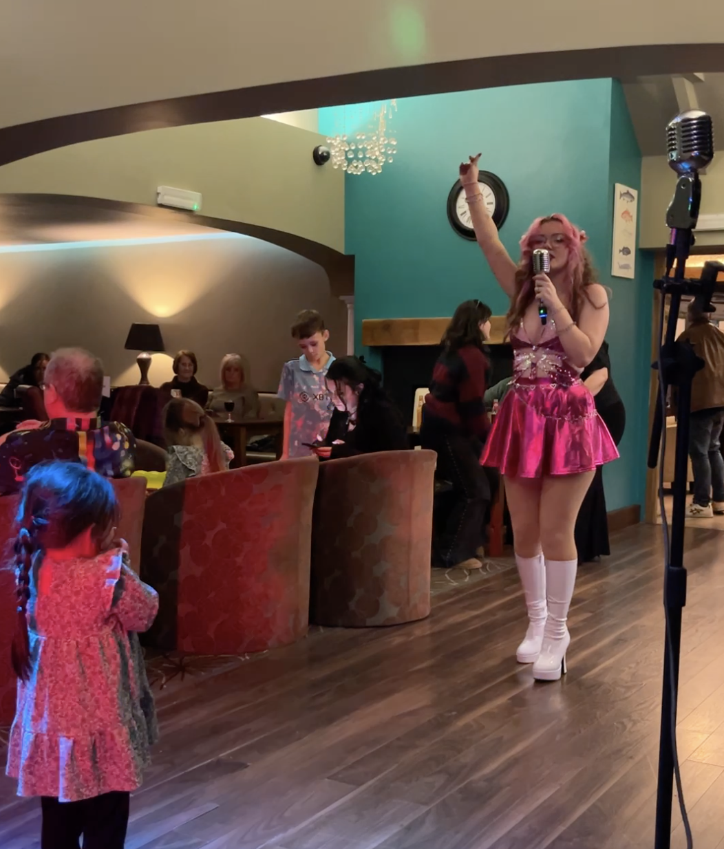 A woman dressed in a pink, shiny, retro-style outfit with white thigh-high boots and pink hair is singing into a microphone on a stage in a restaurant or cafe. She is gesturing upwards with one hand, performing for children and adults seated at tables, with some looking on and children watching attentively.