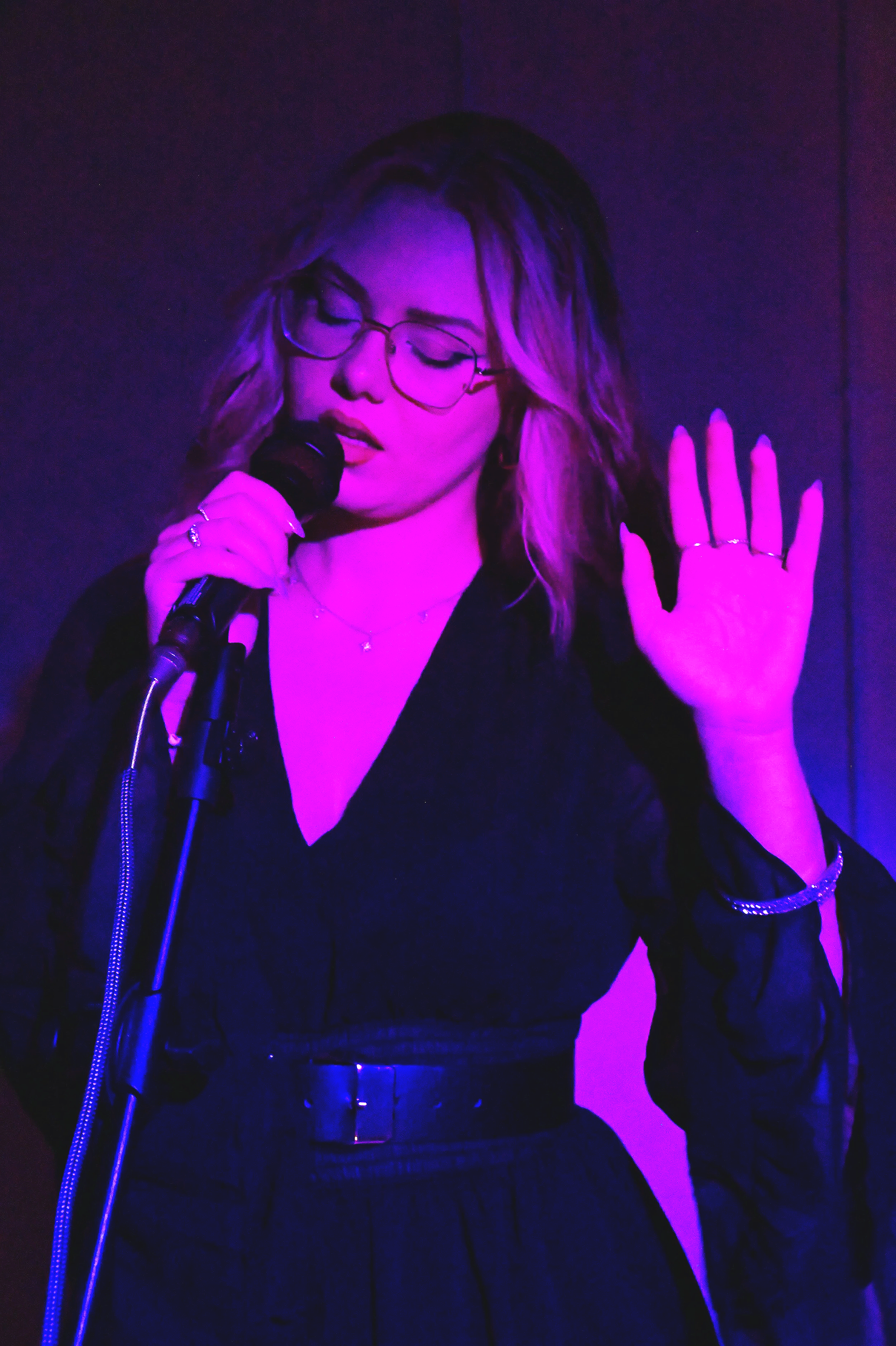 Tay, a woman singing into a microphone with her eyes closed and hand raised expressively at a wedding. She is wearing a black v-neck dress and is illuminated by a dramatic purple stage light.