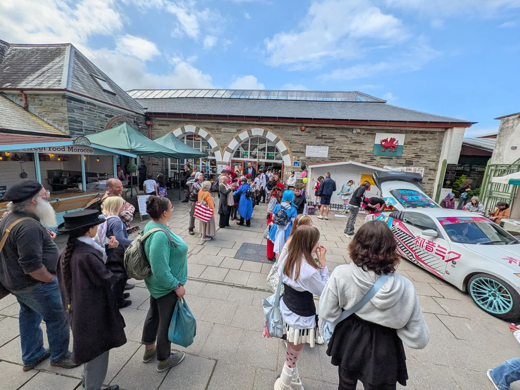 Crowd of people gathered outside a historic stone building with arched windows and Welsh dragon crest, some wearing costumes and colorful clothing, with a racing car parked nearby.