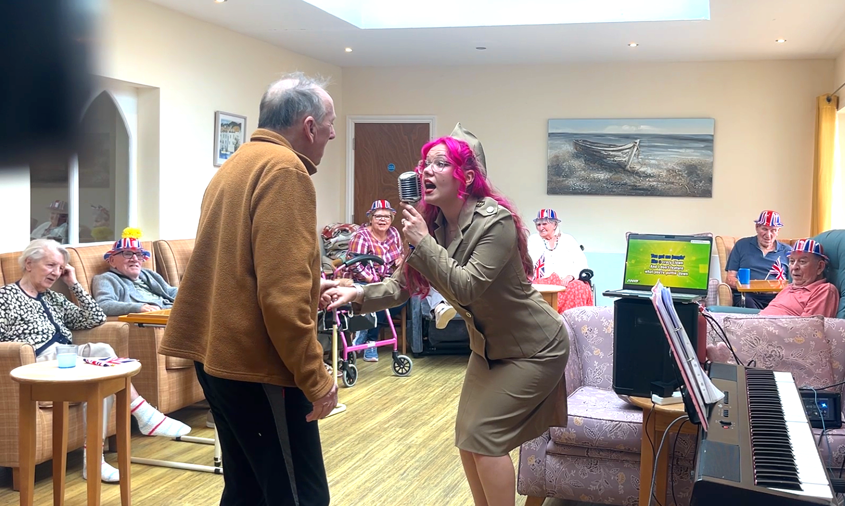 A woman with pink hair singing into a microphone while holding hands with a man in a brown jacket in a room with elderly people. The elderly are seated on sofas and chairs, some wearing British flag hats. A laptop and keyboard are visible near the woman. The room is decorated with maritime artwork and has a cozy, lively atmosphere.
