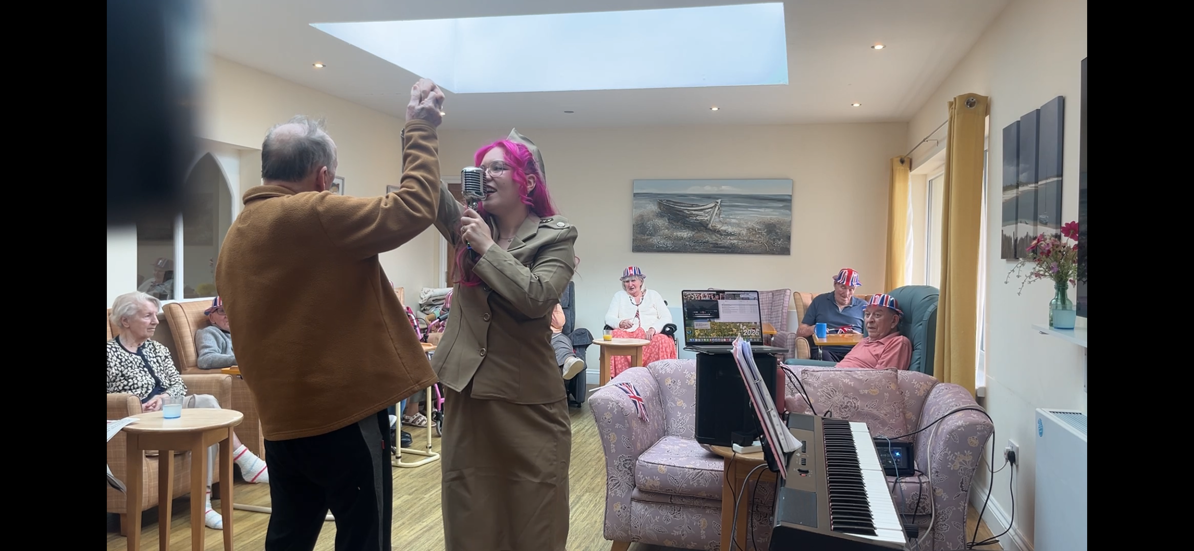 A woman with bright pink hair and glasses singing into a vintage microphone while a man in a brown jacket dances with her. Several elderly people are sitting in a cozy room decorated with yellow curtains, paintings, and Union Jack hats, some watching the performance. A keyboard is on a stand in the foreground.