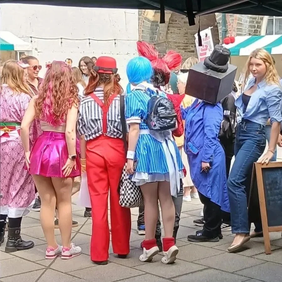 Group of people at a street festival, many dressed in colorful costumes and wigs, with some wearing themed clothing and accessories.