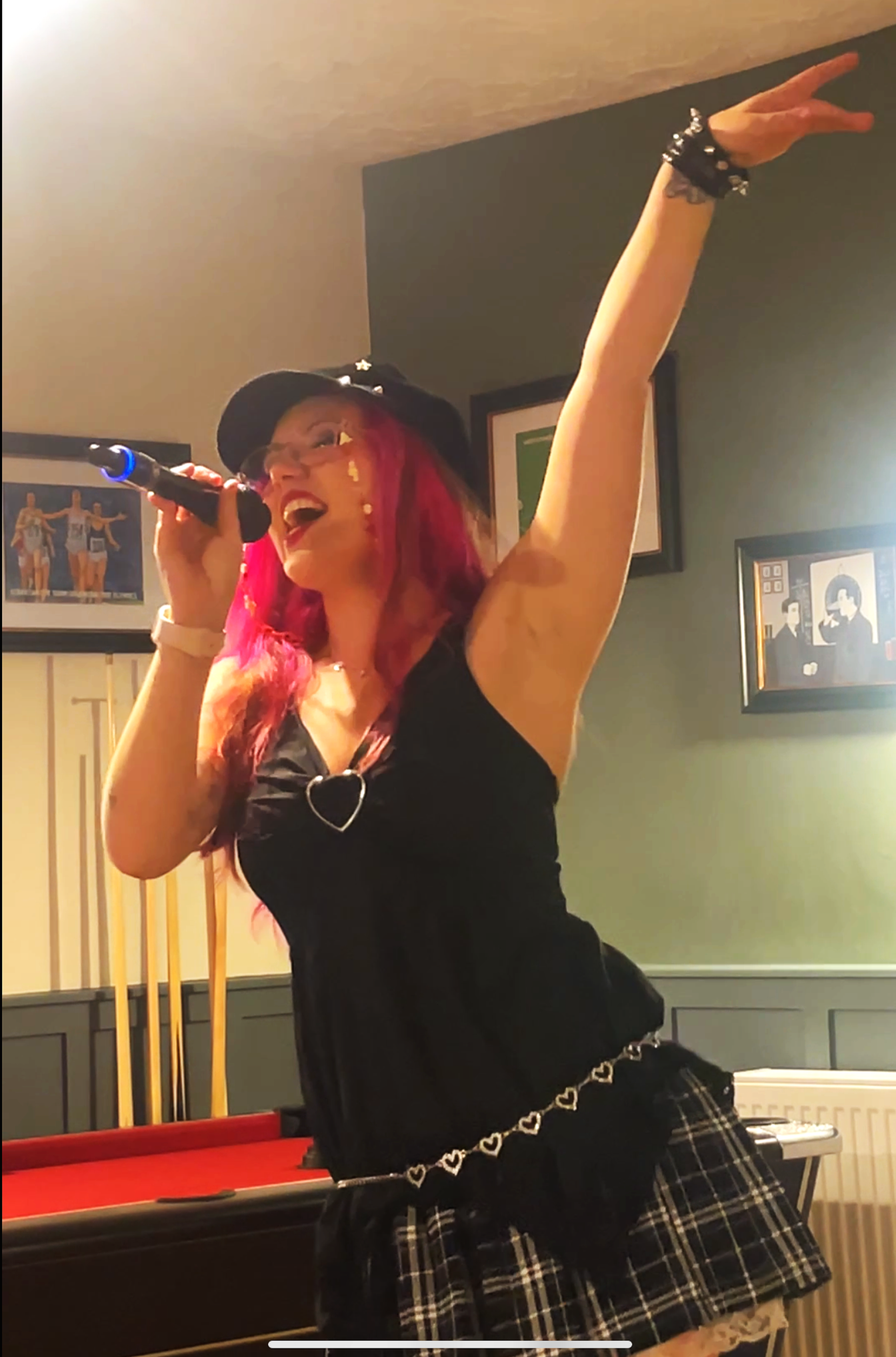 Tay performing in a pub, singing into a microphone with her arm raised high expressively. Her eyes are closed and she is smiling. She wears a black paperboy cap, black v neck dress with a black plaid skirt and a sparkly heart belt. Singing karaoke