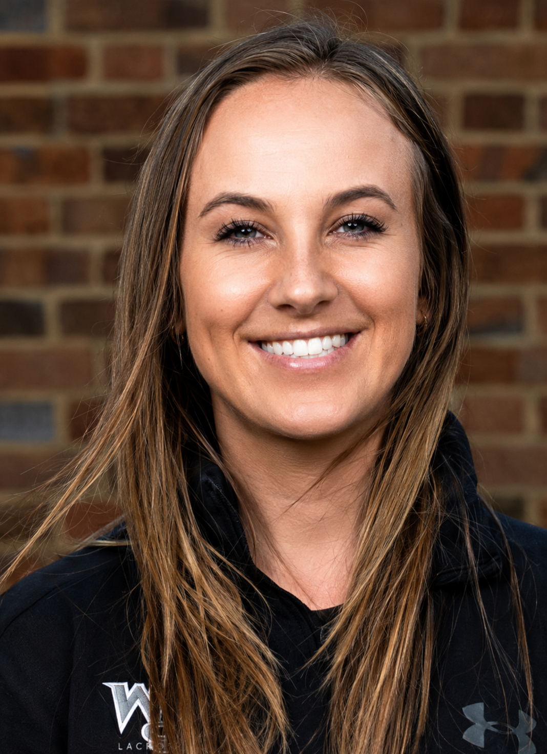 Close-up of a smiling young woman with long brown hair, wearing a black athletic shirt, standing in front of a brick wall.