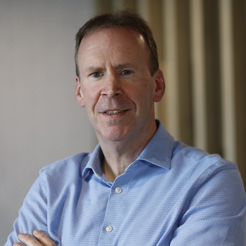Portrait of a middle-aged man with short brown hair, wearing a light blue collared shirt, smiling with arms crossed indoors.