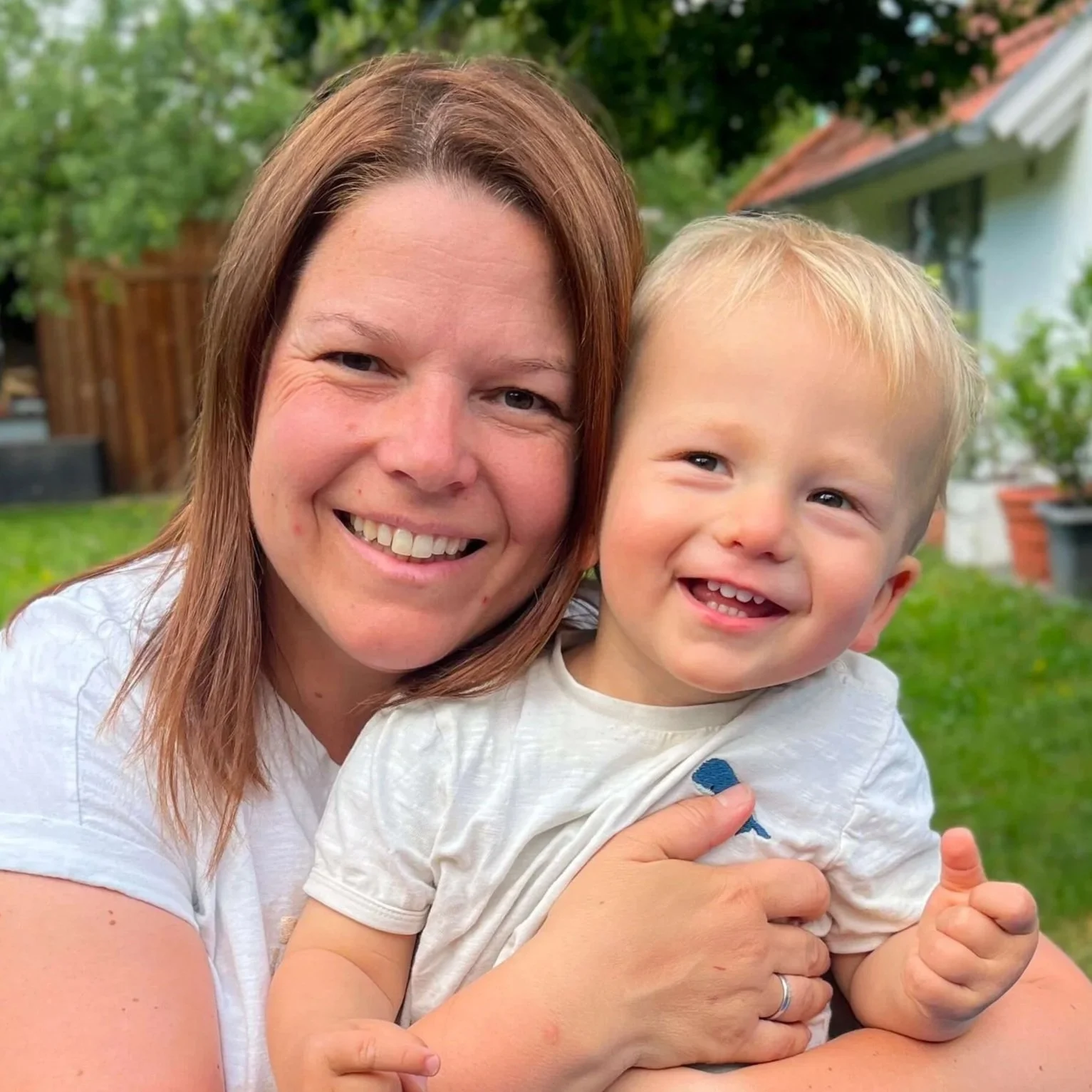 Eine lachende Frau hält einen lachenden kleinen Jungen im Garten, im Hintergrund Bäume und ein Gartenhaus.