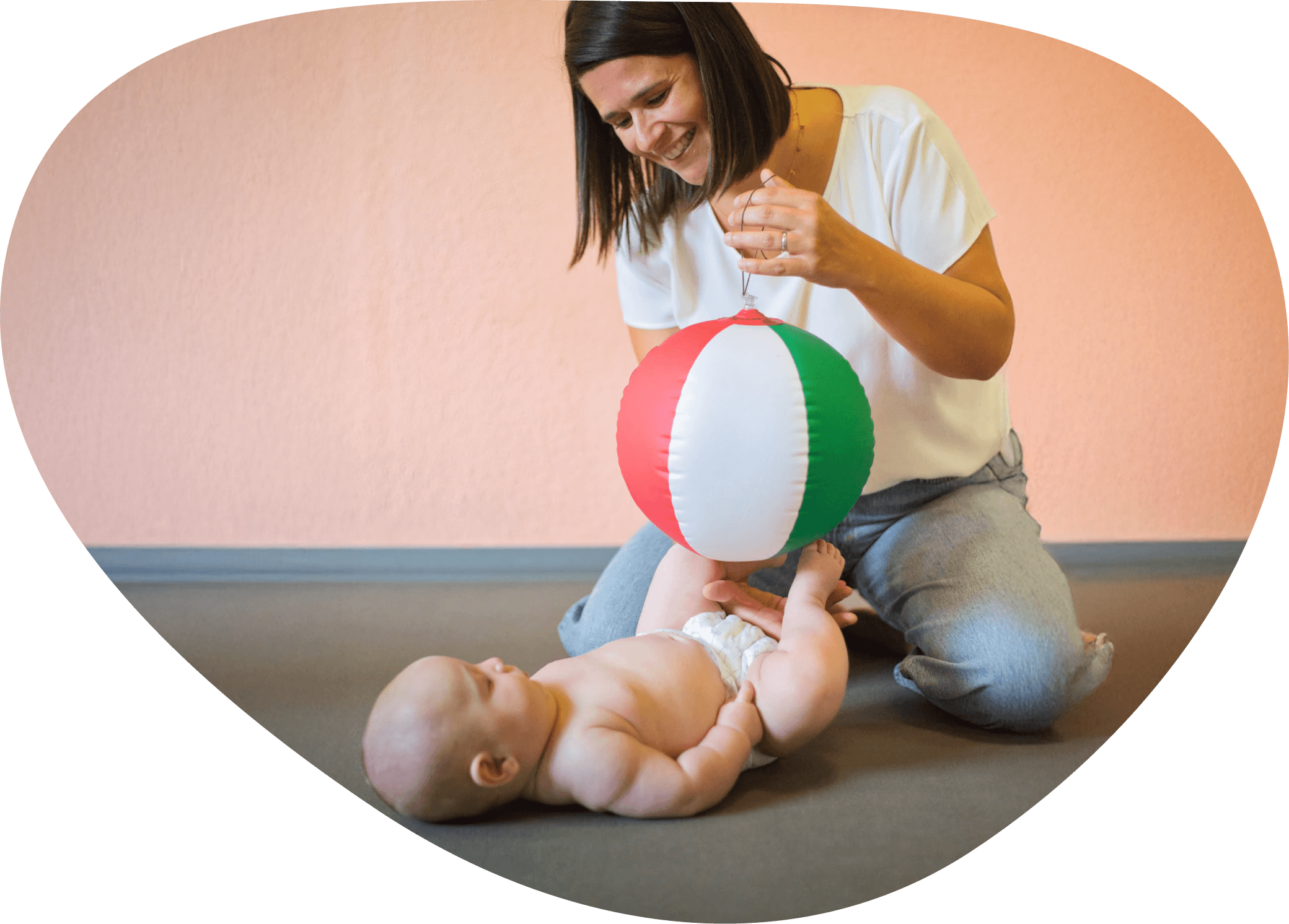 Eine Frau spielt mit einem Baby, das auf dem Boden liegt, während dieses einen bunte Strandball mit den Füßen hält.