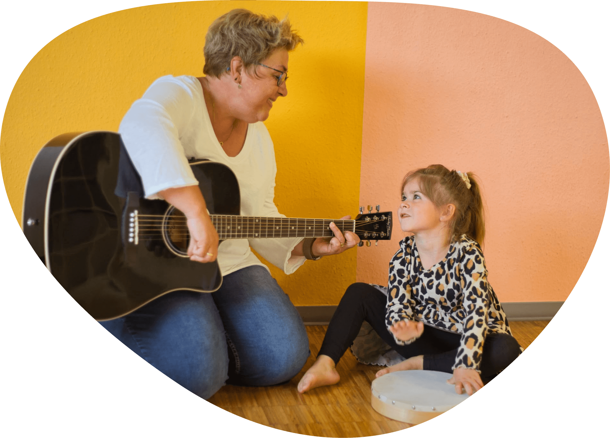 Eine ältere Frau mit kurzen, blonden Haaren und Brille spielt Gitarre, während ein kleines Mädchen mit langem, hellbraunem Haar und Leo-Muster-Shirt ein Tamburin festhält. Sie sitzen im Raum vor einer Wand, die halb gelb und halb rosafarben ist.