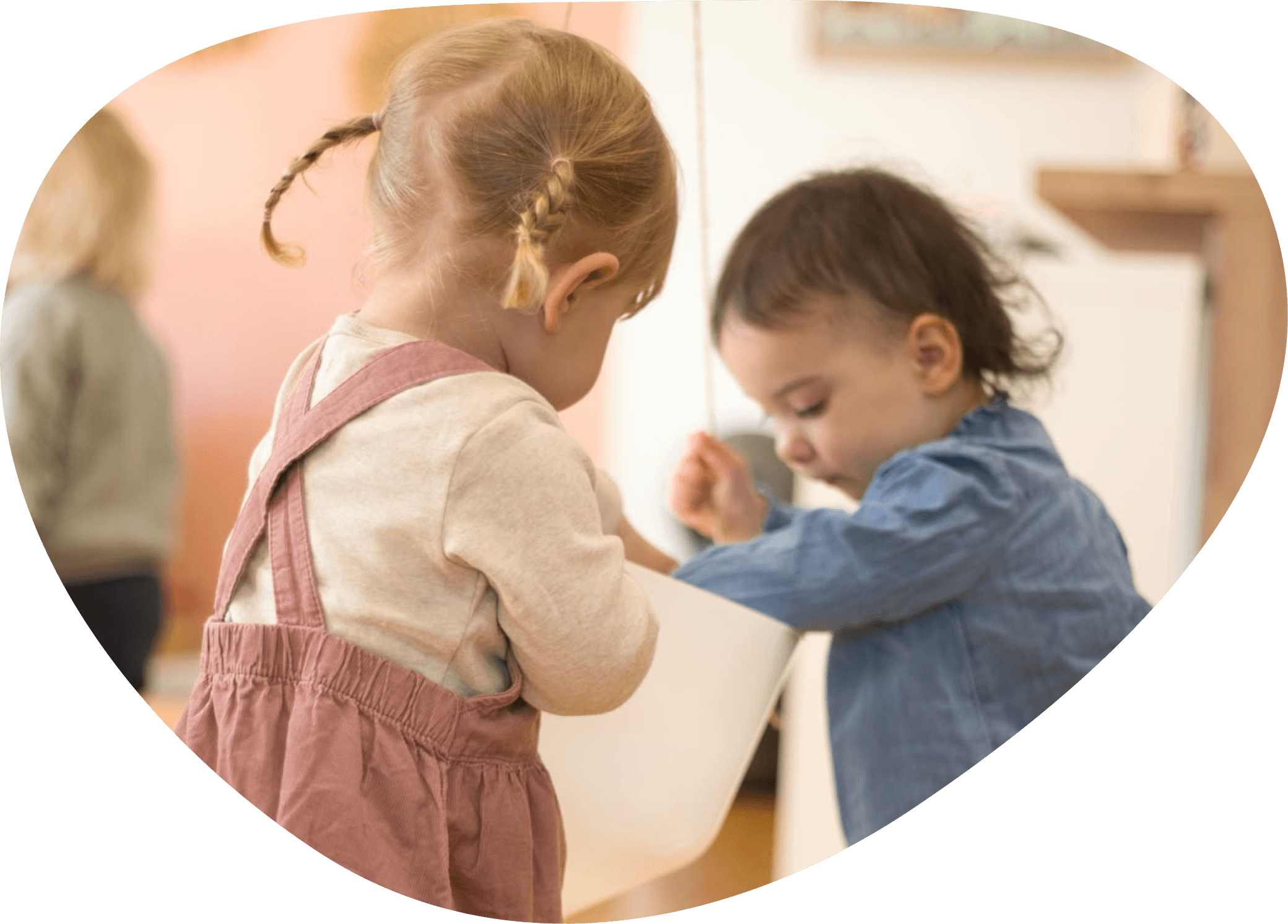 Zwei Kinder, ein Mädchen mit blonden geflochtentn Zöpfen und ein Junge mit dunklen Haaren, spielen zusammen, im Hintergrund sind weitere Kinder.