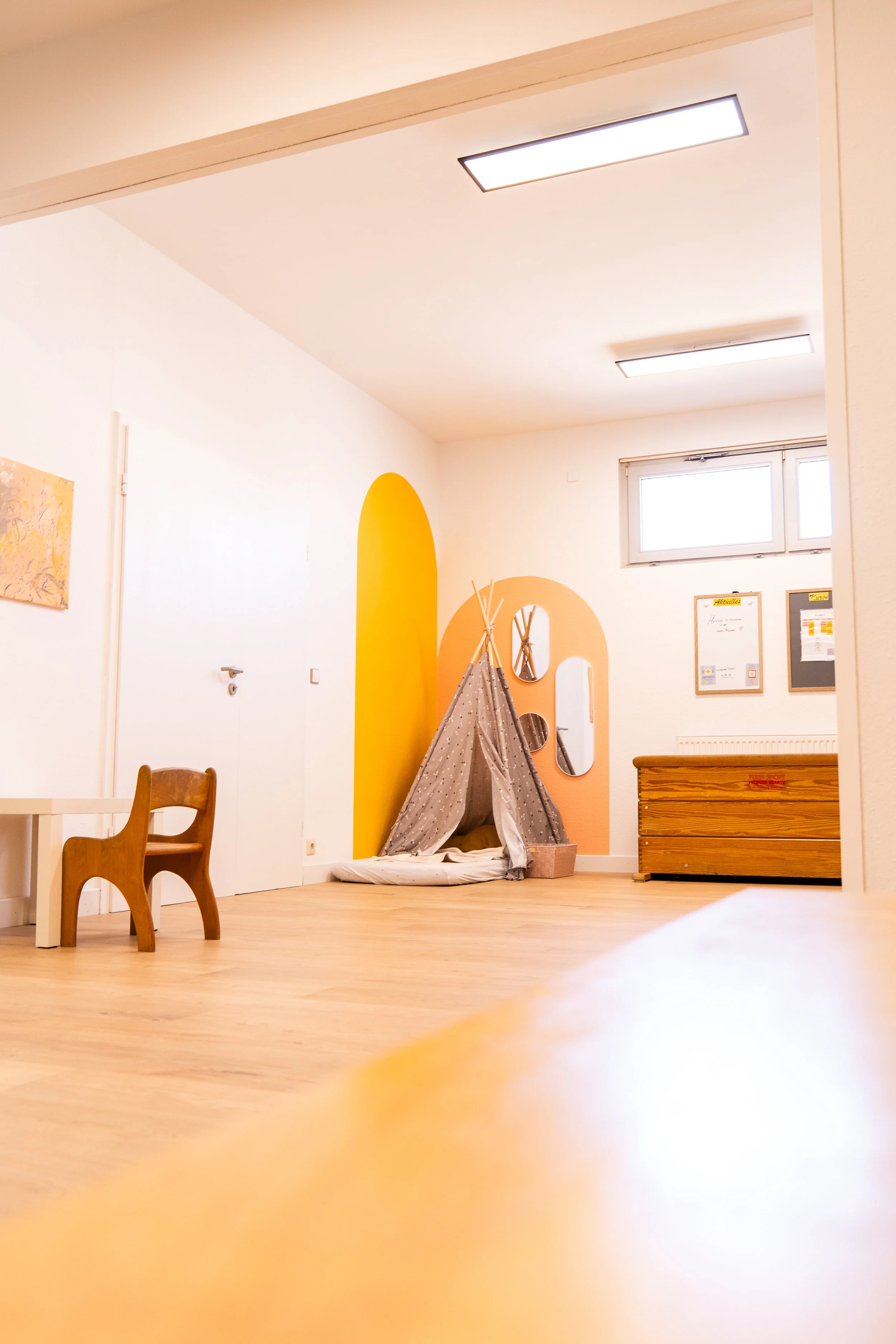 Ein Kinderzimmer mit weißen Wänden, gelber Akzentwand, einem kleinen Zelt, einem Stuhl und einem Regal. Es gibt einen Fenster und einige eingerahmte Dokumente an der Wand.