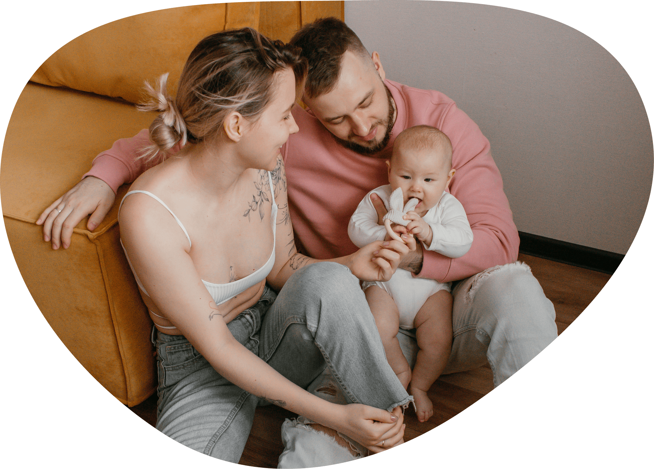 Eine vierköpfige Familie sitzt auf einem Sofa, lacht und genießt die Zeit miteinander. Die Mutter hat braune Haare, trägt ein weißes Top und hat eine Tätowierung am Arm. Der Vater trägt einen pinkfarbenen Pullover, hat braune Haare und einen Bart. Das Baby hat kurze, helle Haare, trägt ein weißes Outfit und hält eine Schnullerkette im Mund. Sie scheinen eine glückliche Familie zu sein, die gemeinsam Zeit verbringt.