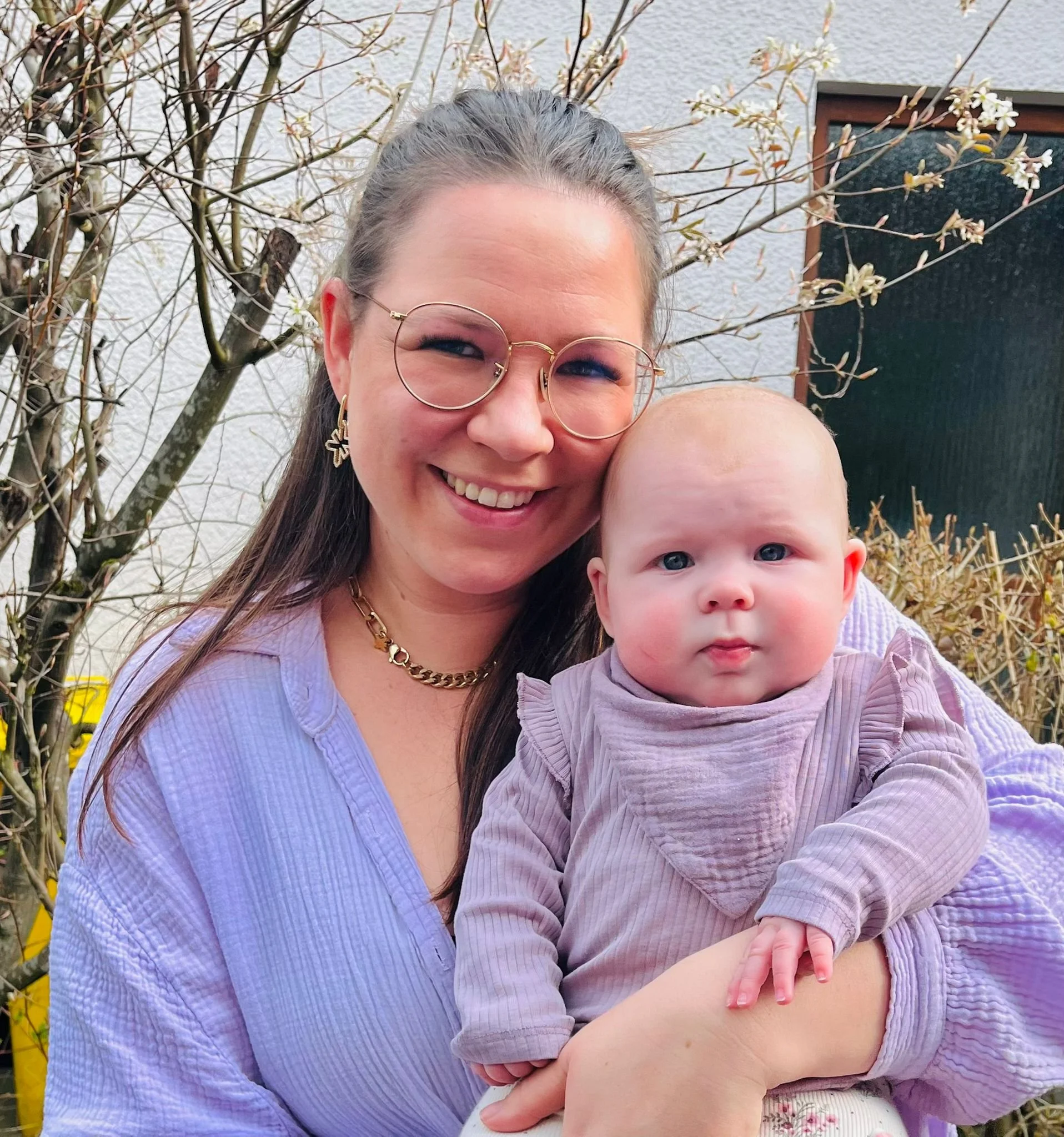 Nahaufnahme von einer lachenden Frau mit Brille und einem Baby, die im Garten vor einer weißen Wand und einer blühenden Pflanze fotografiert wurden.