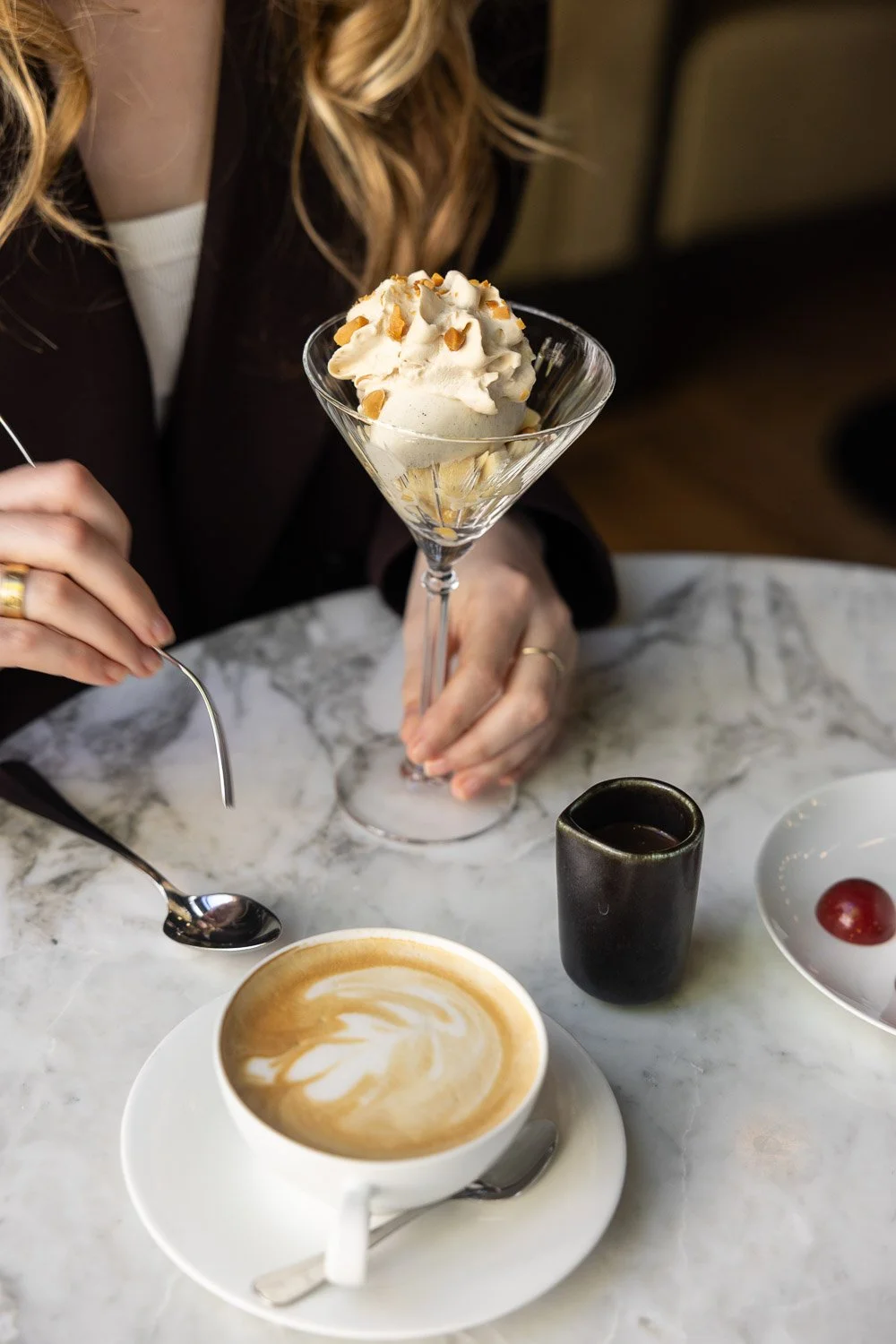 Een persoon houdt een cocktailglas met vanille-ijs versierd met noten. Op de tafel staan een kopje koffie met latte art, een klein glas met een drankje, en een wit schaaltje met een druif.