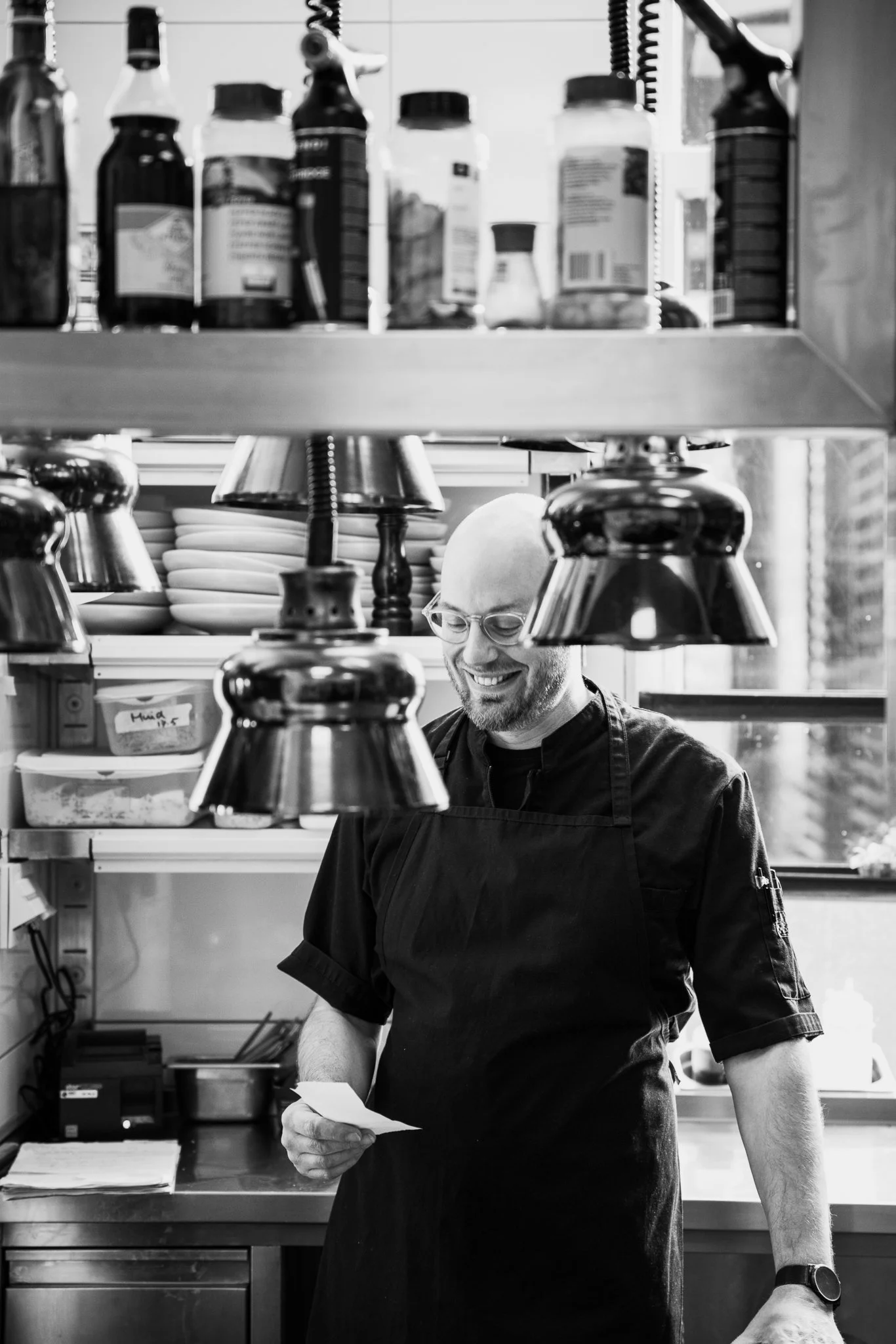 Man in een zwarte kokspak met een bril leest een stuk papier in een keuken met plateaus en schappen vol potten en bestek.