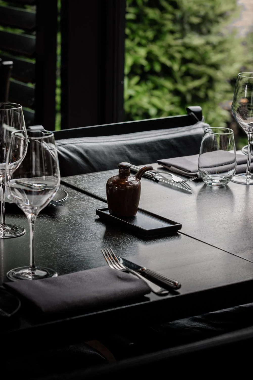 Een gedekte dinertafel met wijnglazen, bestek, een grijze servet, een donkere tafel en een groengeblokkeerde achtergrond.